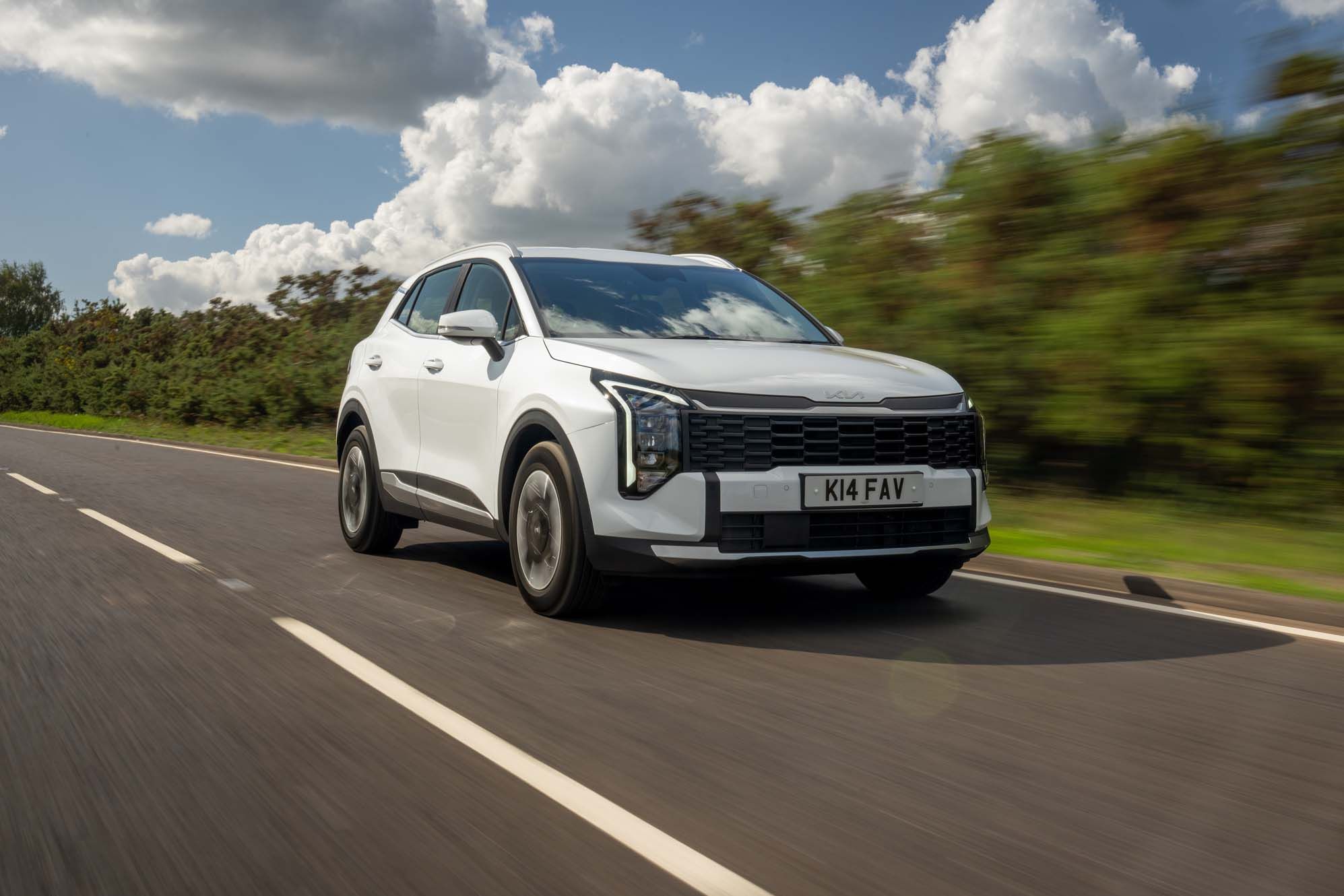 White Kia Sportage driving on a rural road with greenery in the background under a partly cloudy sky.
