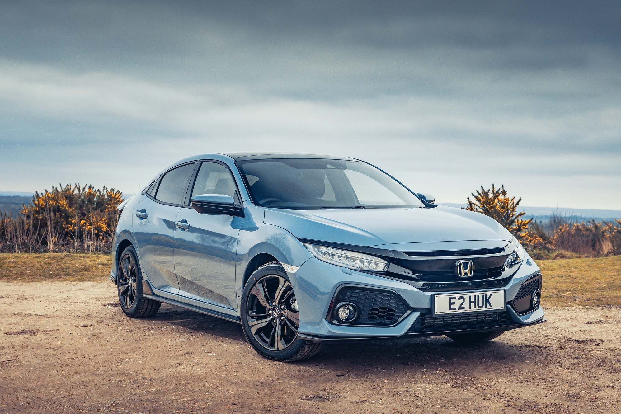 A grey Honda Civic hatchback parked on a dirt road, under a cloudy sky, with distant hills and sparse shrubs in the background.