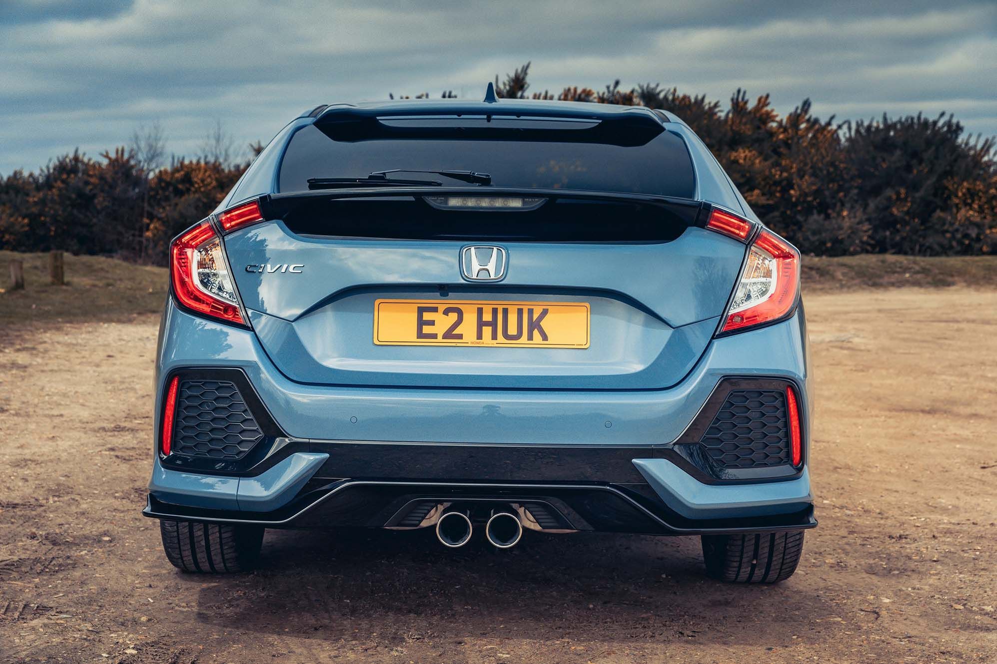 Rear view of a grey Honda Civic hatchback parked on a dirt road, featuring a dual exhaust and a yellow license plate.