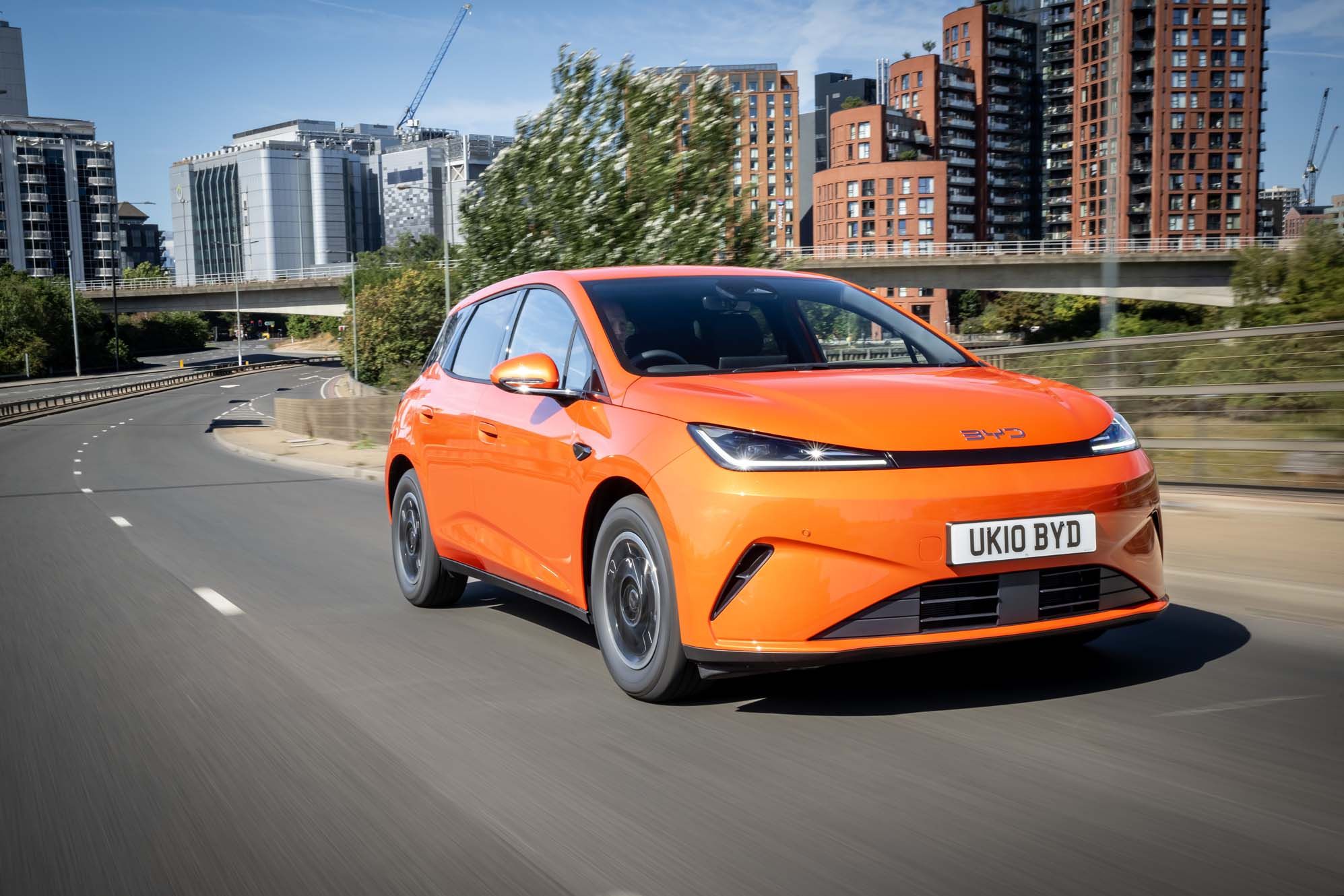 Orange BYD DOLPHIN G DM-i driving on a city road with modern buildings and greenery in the background.