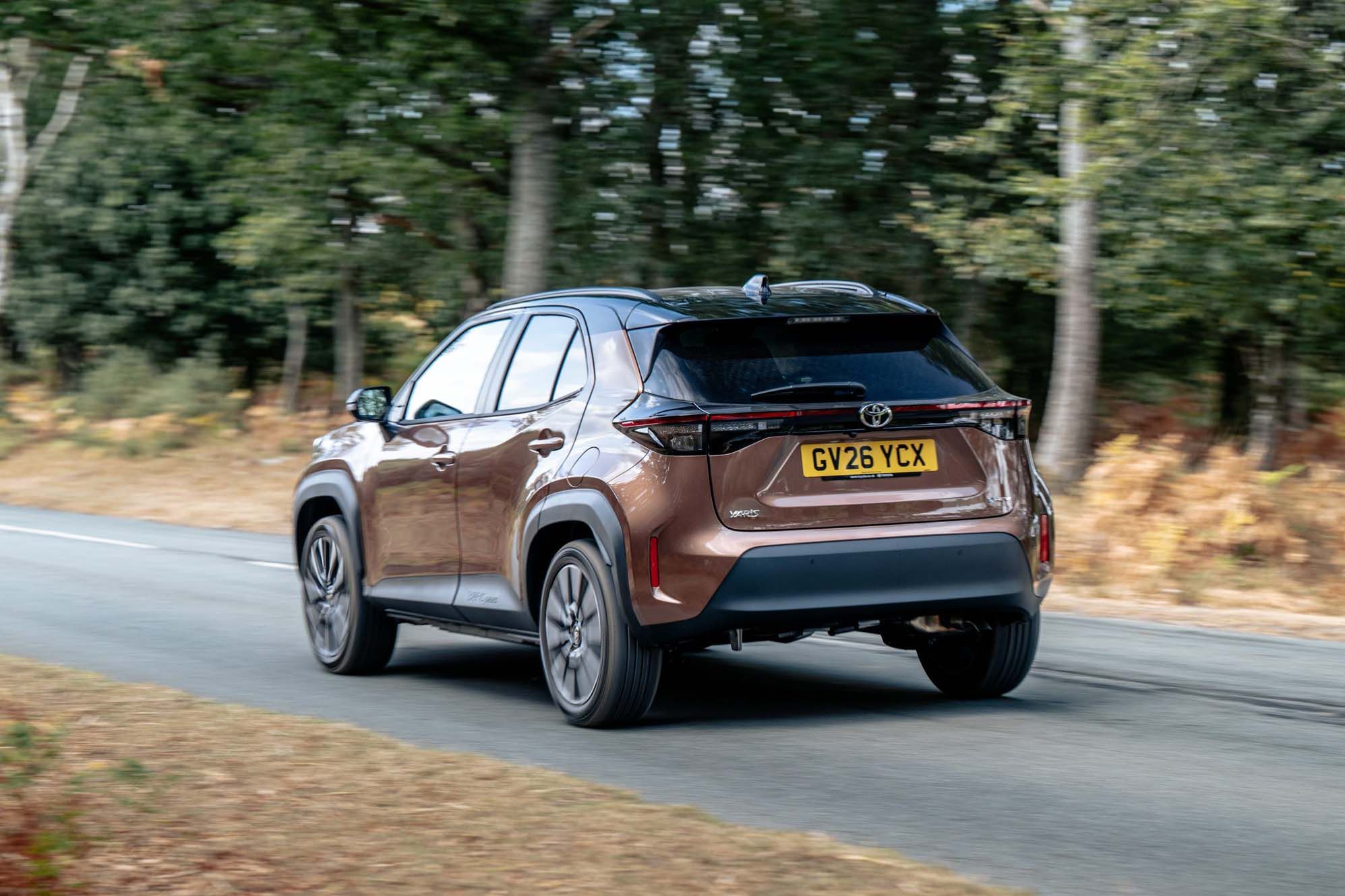 A bronze Toyota Yaris Cross with a UK license plate driving along a tree-lined road.