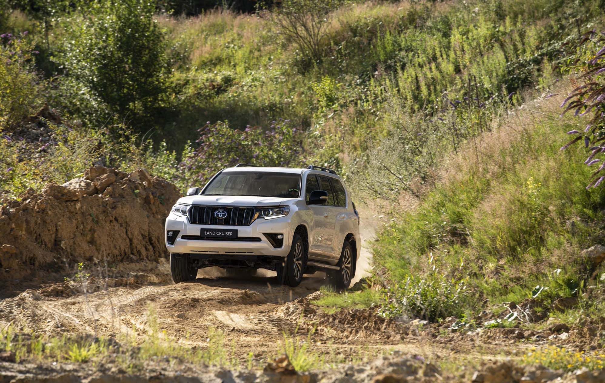 A white Toyota Land Cruiser navigates a rugged dirt path surrounded by green vegetation and hilly terrain.