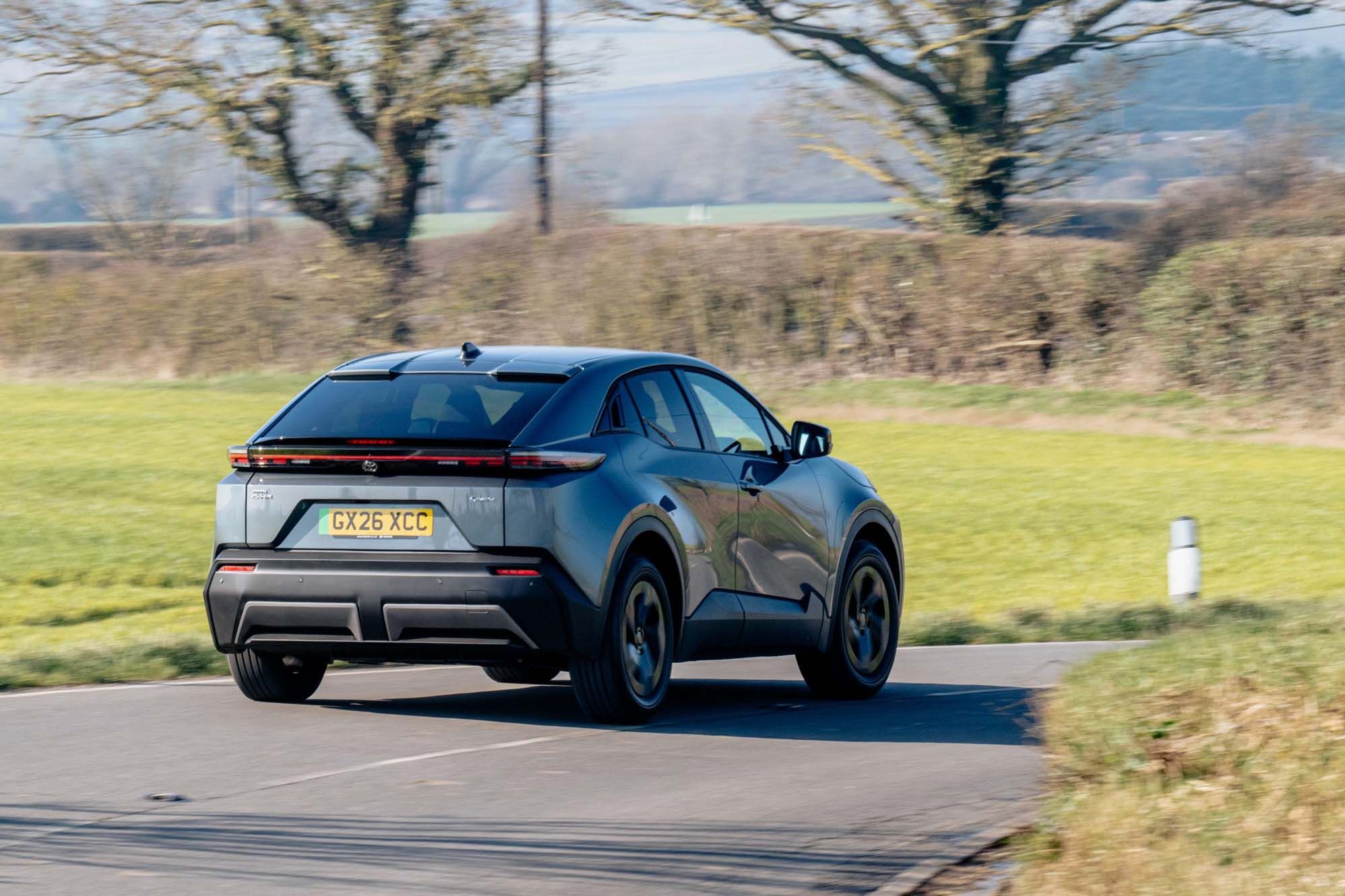 Grey Toyota C-HR+ with number plate GX26 XCC drives on a rural road, surrounded by green fields and trees under a clear blue sky.