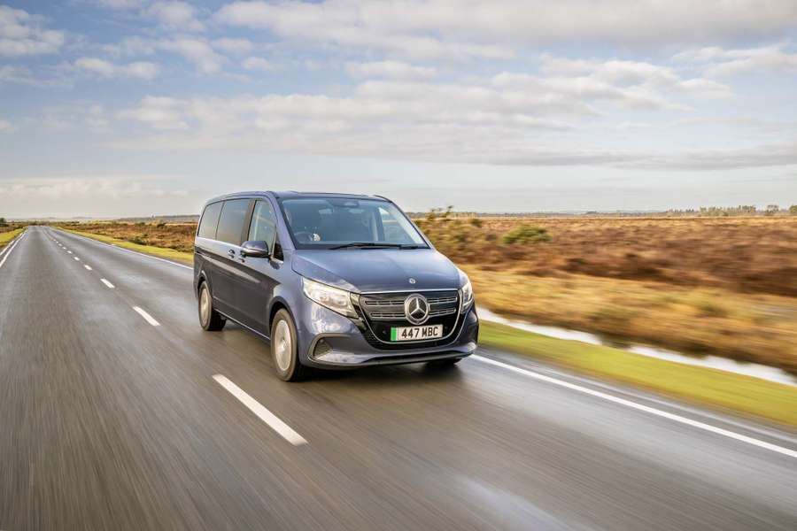 A grey Mercedes EQV driving down a straight, open road in a rural landscape under a partly cloudy sky.