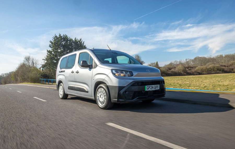 Silver Toyota Proace City Verso driving on a rural road, surrounded by trees and fields, under a clear blue sky.