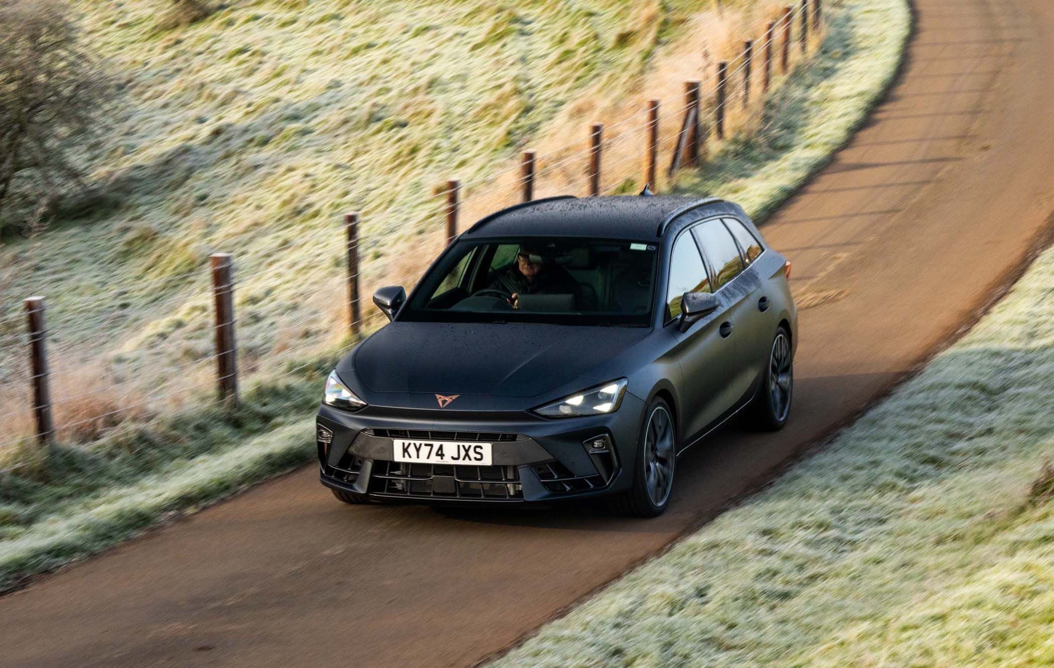 A dark-colored CUPRA Leon Estate drives on a narrow, frosty rural road lined with wooden fences and grass.