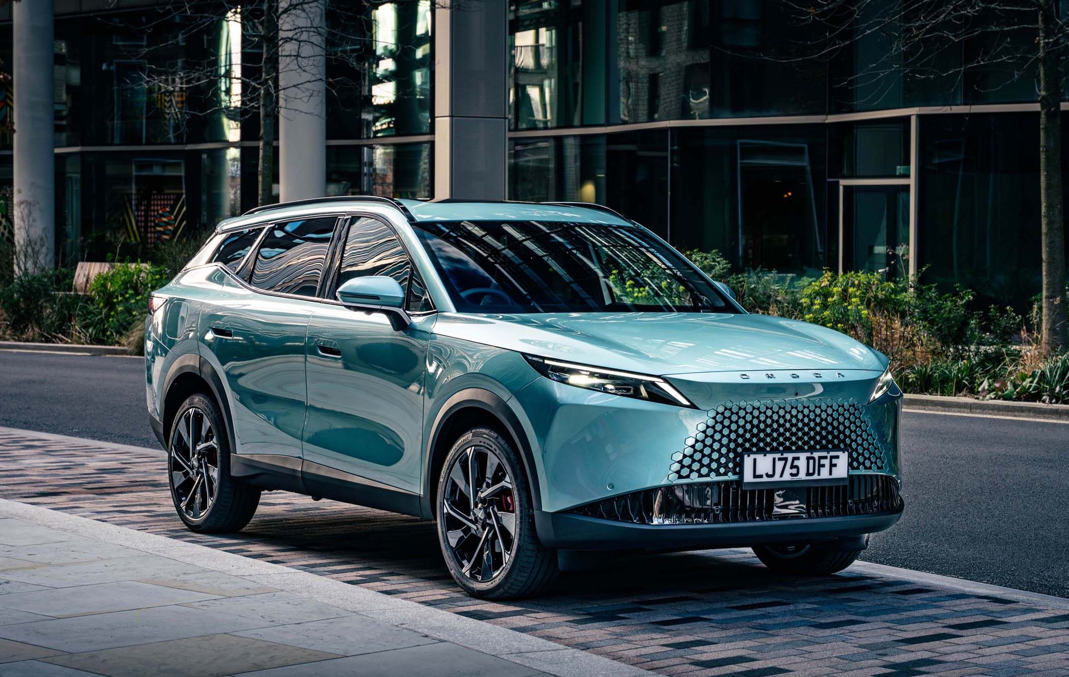 A blue OMODA 7 parked on a city street, featuring sleek design and black wheels, with buildings and greenery in the background.