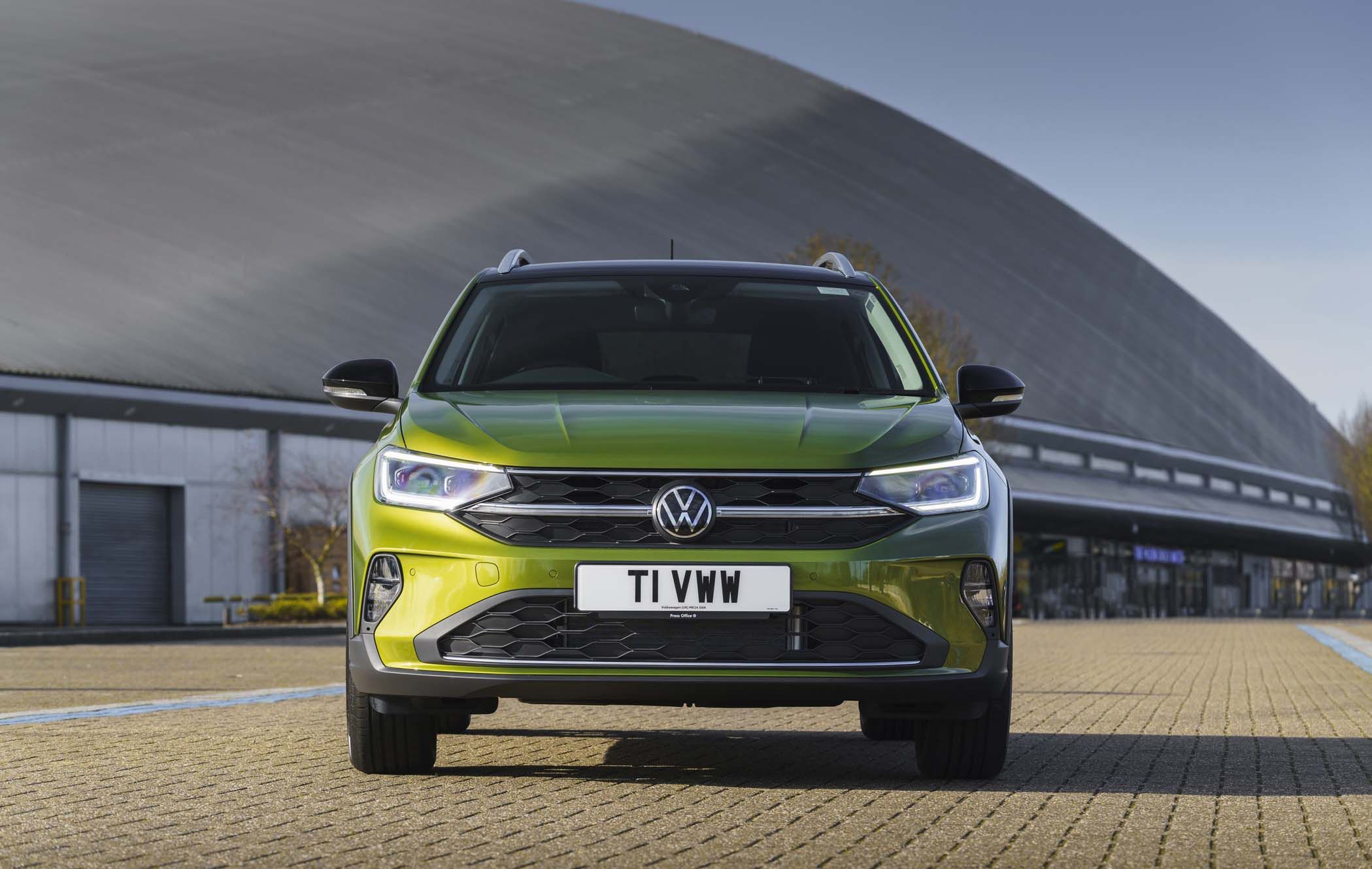 A green Volkswagen Taigo is parked on a paved area in front of a large, curved building under a clear sky.