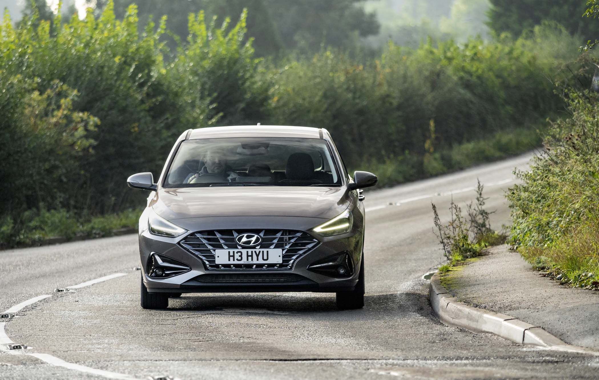 A silver Hyundai i30 drives on a curvy road lined with greenery on a clear day.