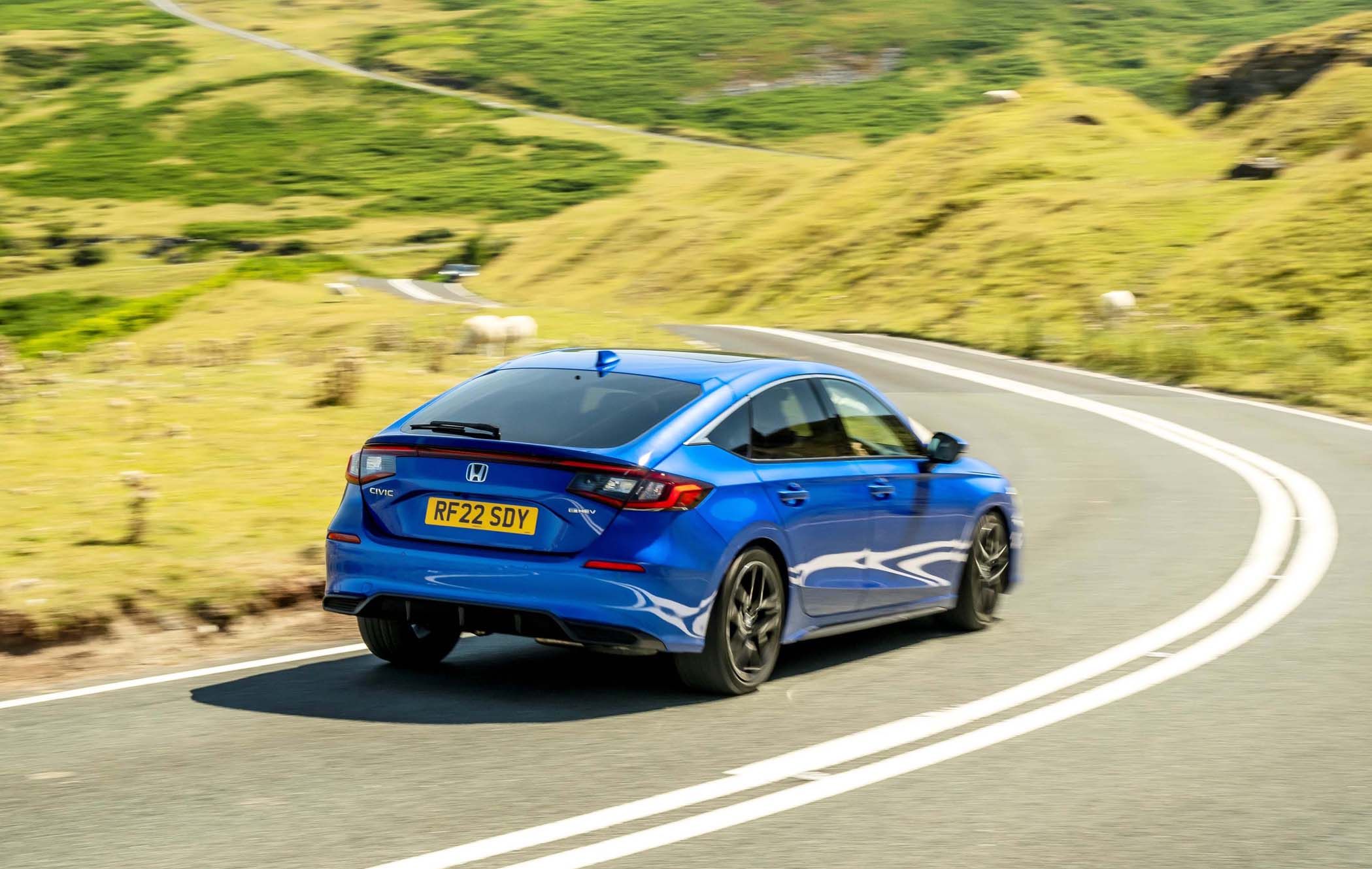 A blue Honda Civic drives along a winding road through lush green hills on a sunny day.
