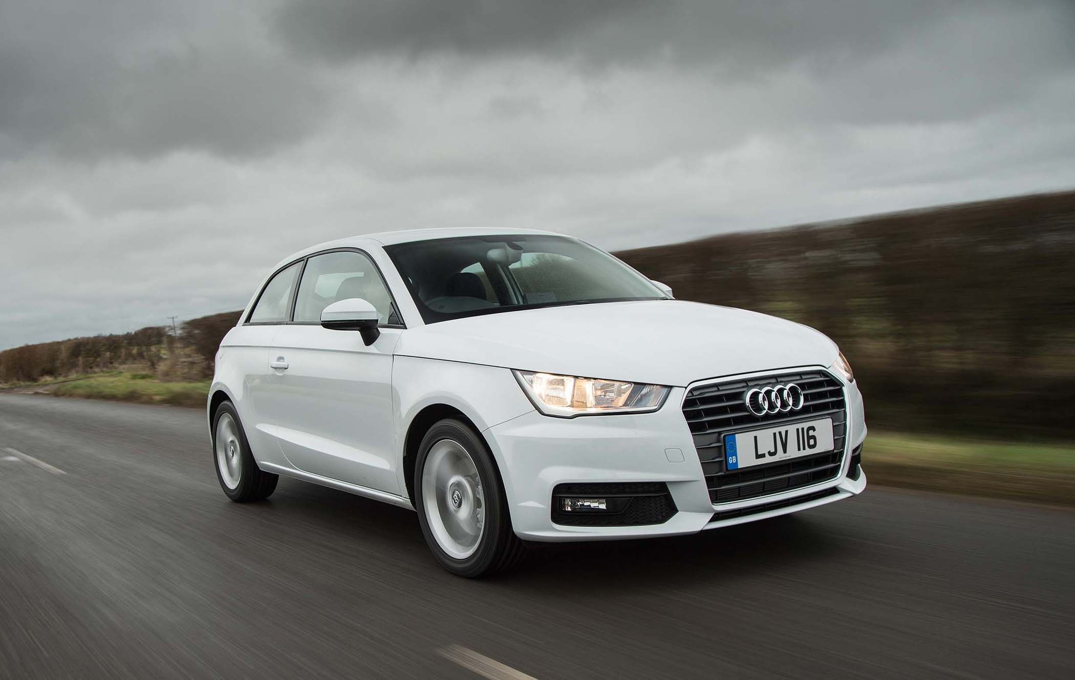 White Audi A1 driving on a road with blurred motion, under a cloudy sky.