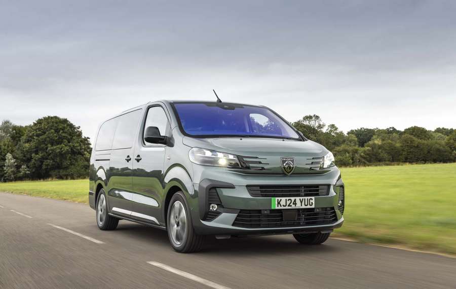 Green van driving on a rural road with fields and trees in the background under a cloudy sky.