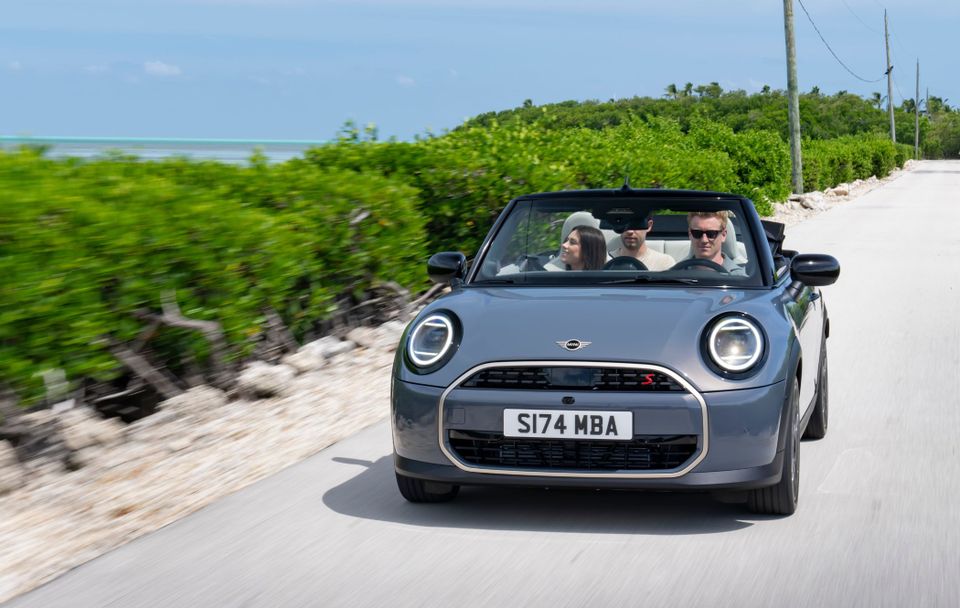 A grey convertible MINI with three passengers drives on a sunny road lined with greenery and a clear blue sky.