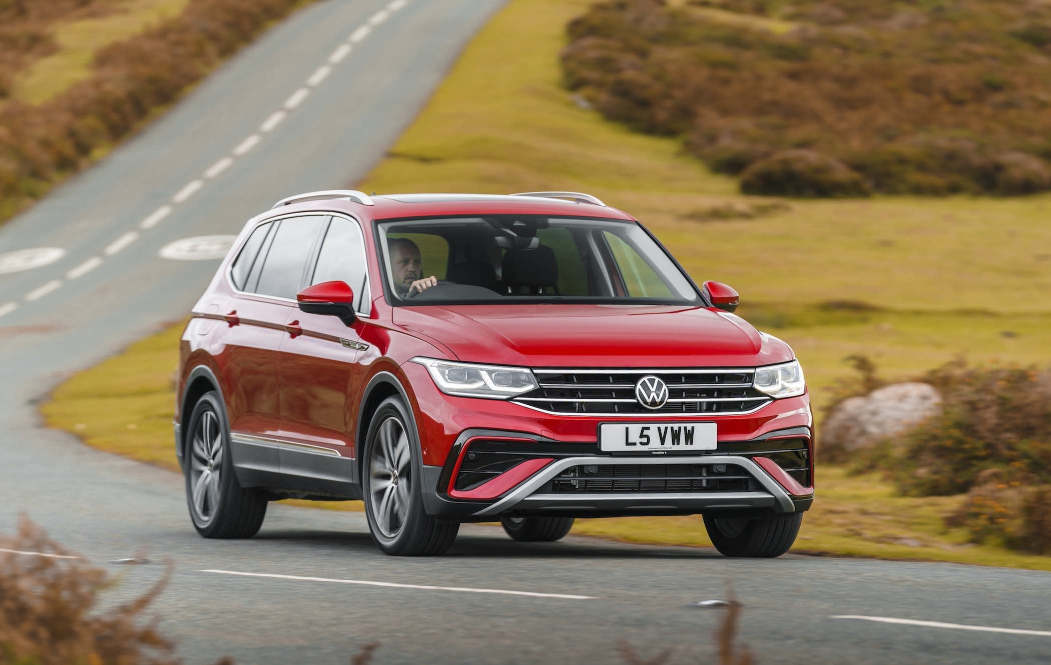 Red Volkswagen Tiguan Allspace driving on a scenic road with grassy hills in the background.