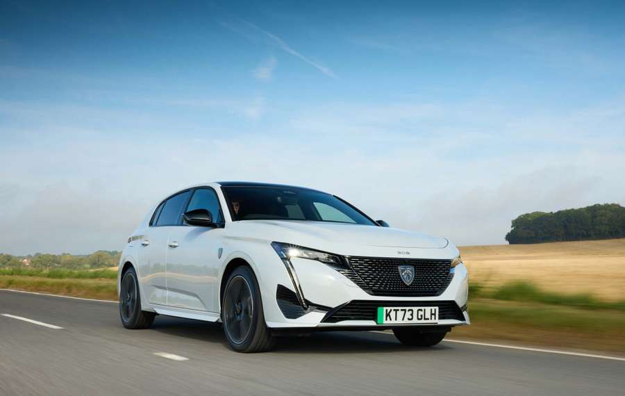 White Peugeot E-308 driving on a rural road with fields and trees in the background under a clear blue sky.