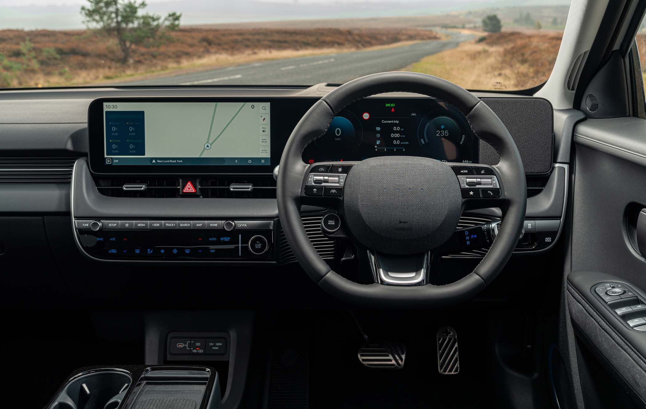 Interior view of a Hyundai IONIQ 5 showing a modern dashboard with a digital display, navigation screen, steering wheel, and control buttons. Road visible ahead.