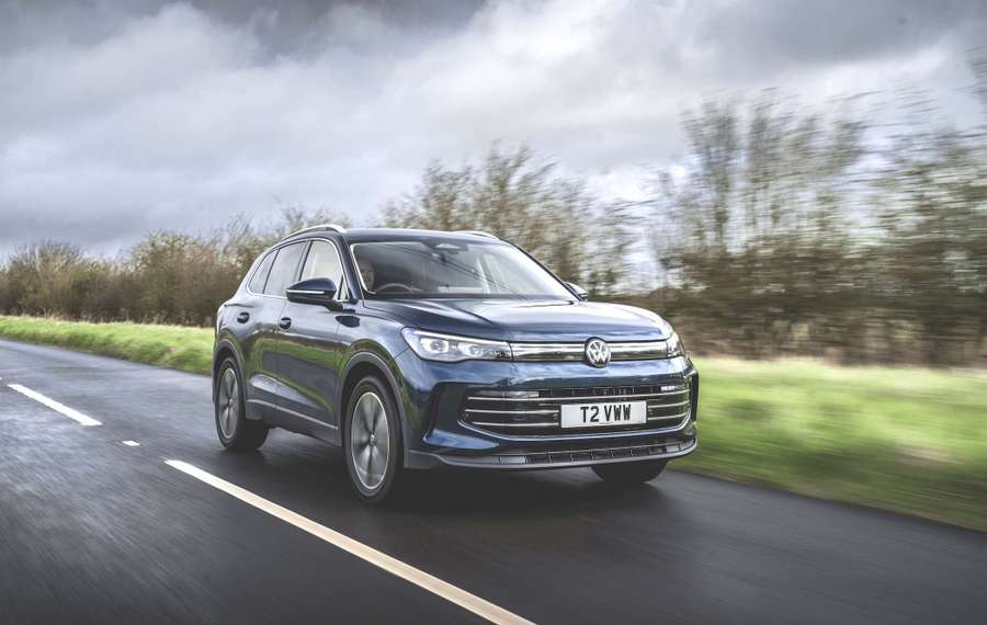 A blue VW Tiguan drives on a rural road with blurred trees in the background under a cloudy sky.
