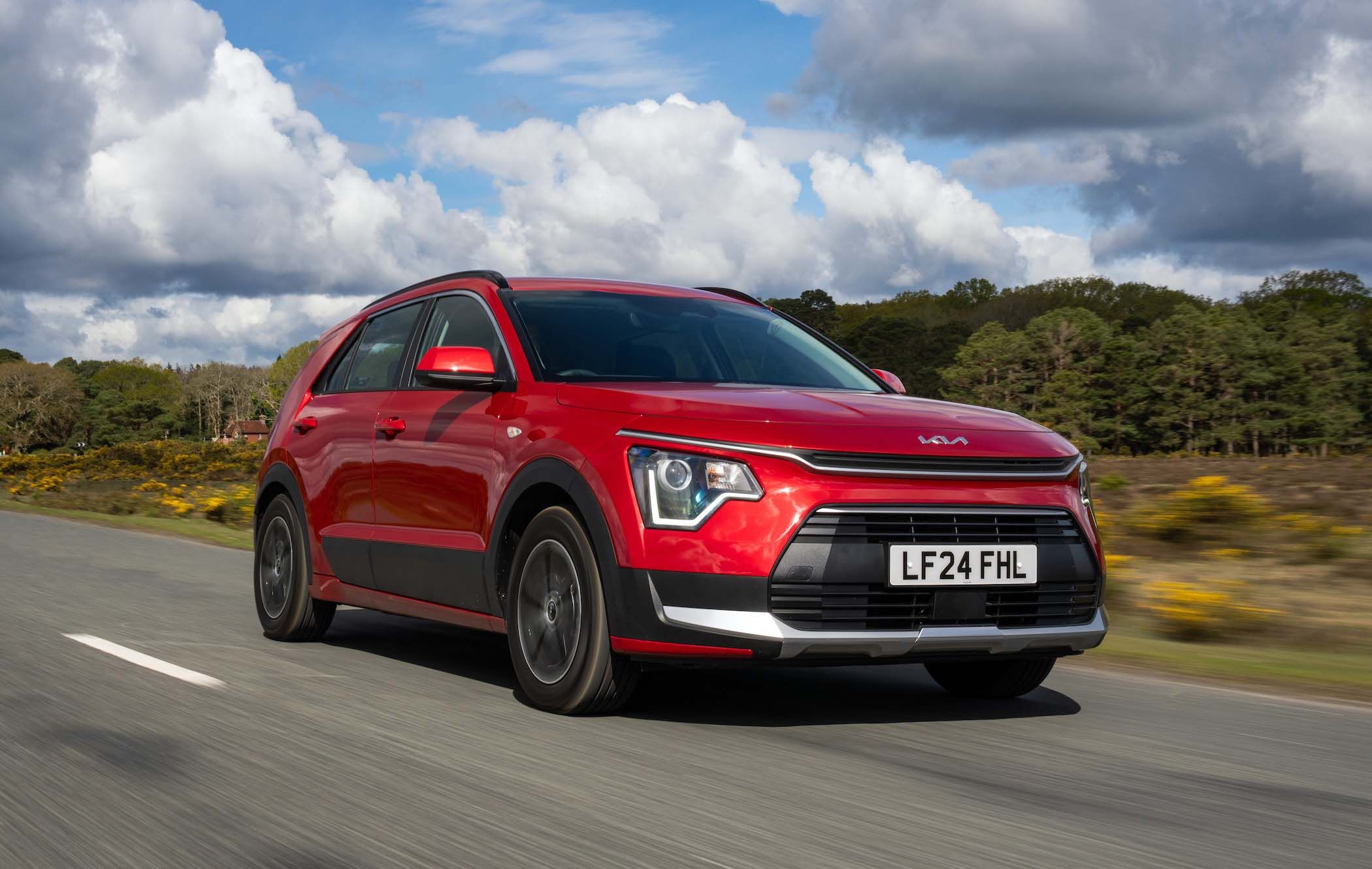 Red Kia Niro driving on a rural road, surrounded by greenery and a cloudy sky.