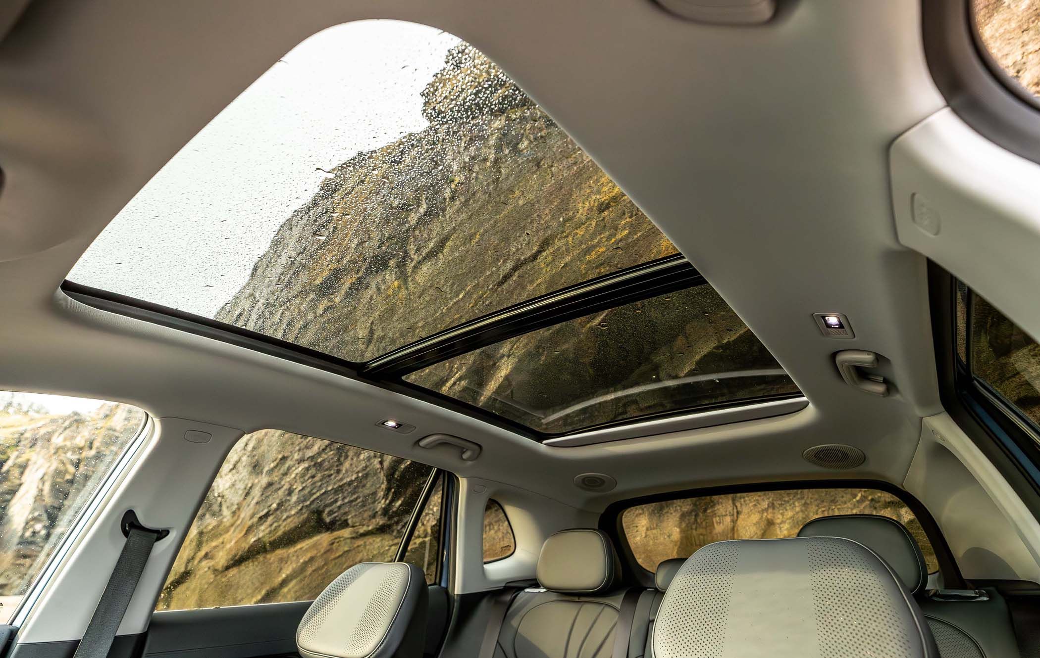 Interior view of a Geely Starray with a panoramic sunroof, showing raindrops on the glass and a rocky cliff background.