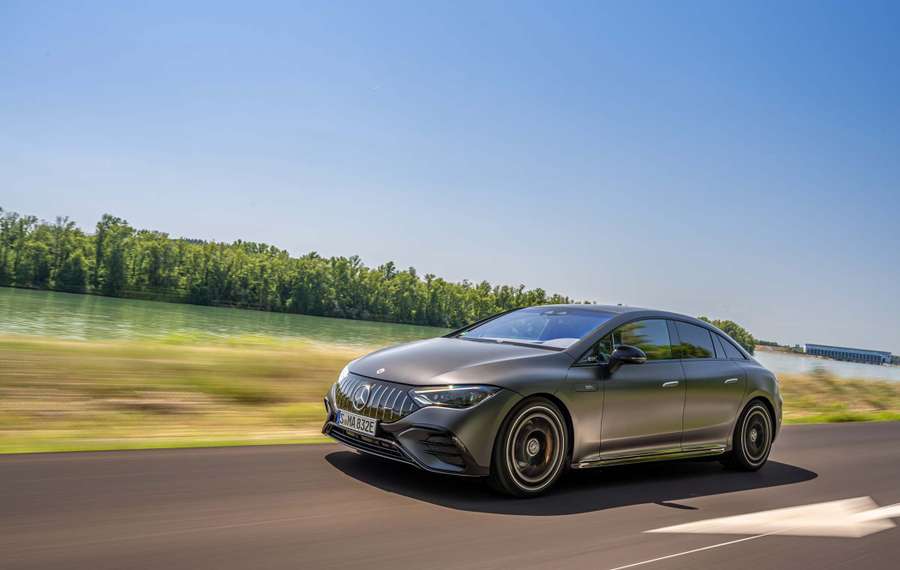 A grey Mercedes-AMG EQE 53+ speeds along a road beside a river, with trees and a clear blue sky in the background.