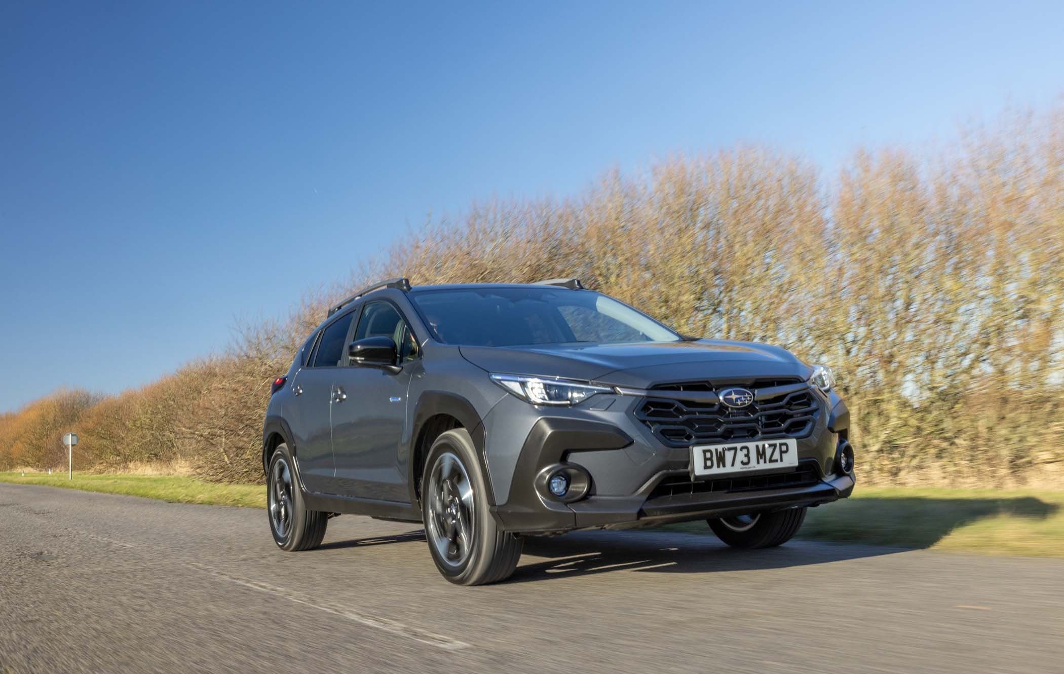 A grey Subaru Crosstrek drives on a rural road under a clear blue sky, with leafless trees lining the roadside.