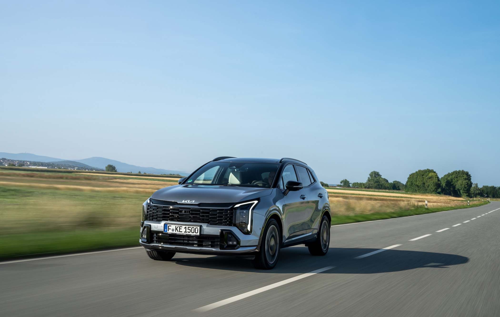 A silver Kia Sportage driving on a rural road with fields and distant hills under a clear blue sky.