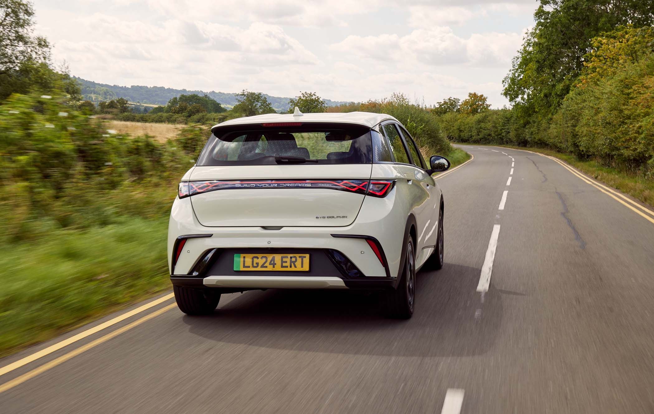 A cream BYD DOLPHIN drives down a scenic, winding rural road bordered by trees and fields under a partly cloudy sky.