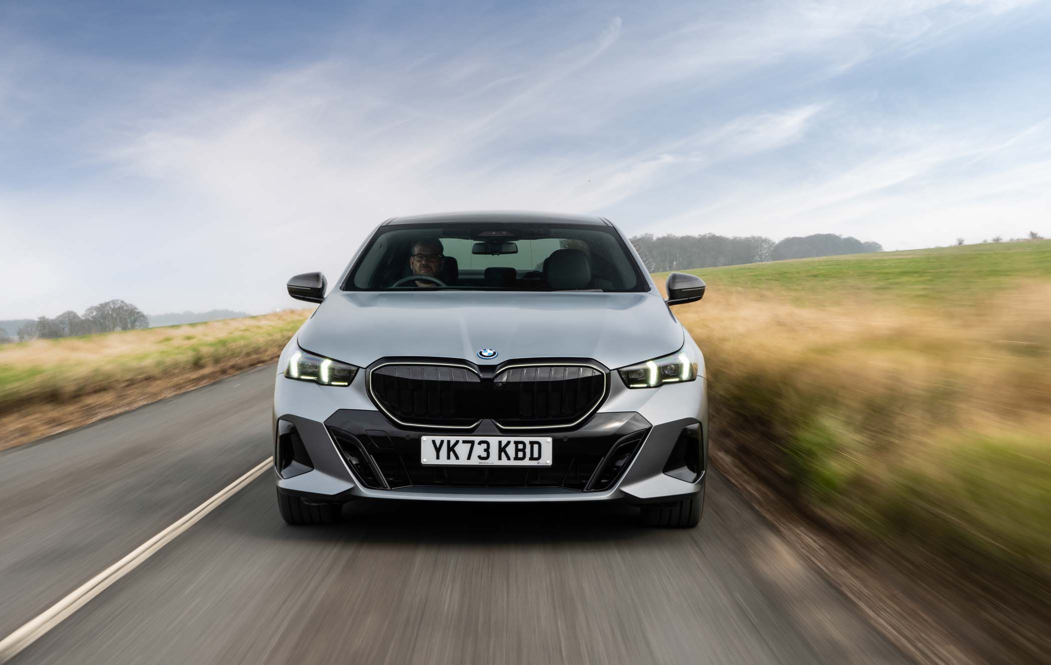 Front view of a silver BMW car driving on a rural road, with blurred motion effect and fields on either side under a clear sky.