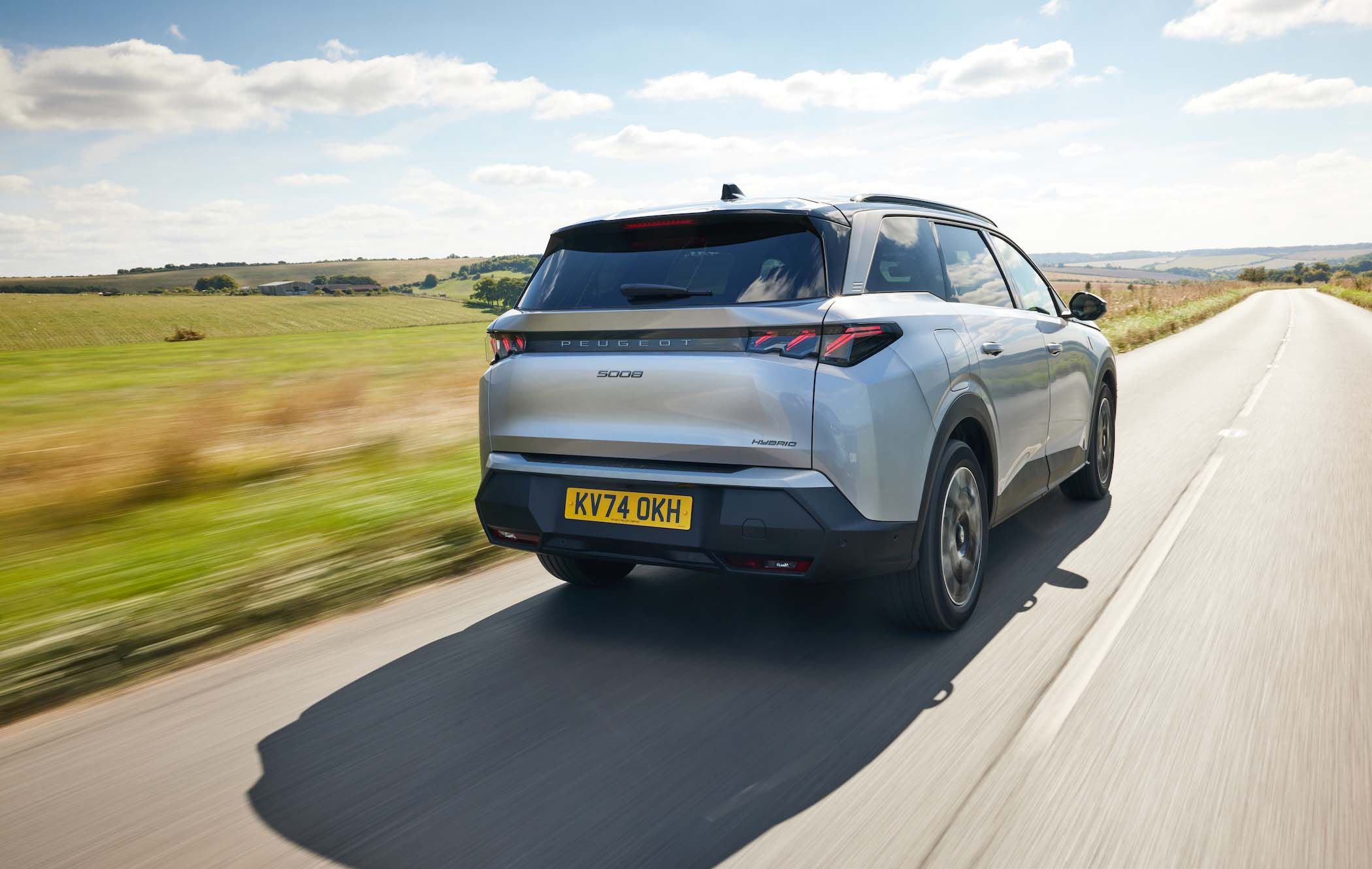 Silver Peugeot 5008 driving on a rural road under a blue sky with clouds, surrounded by fields and hills.