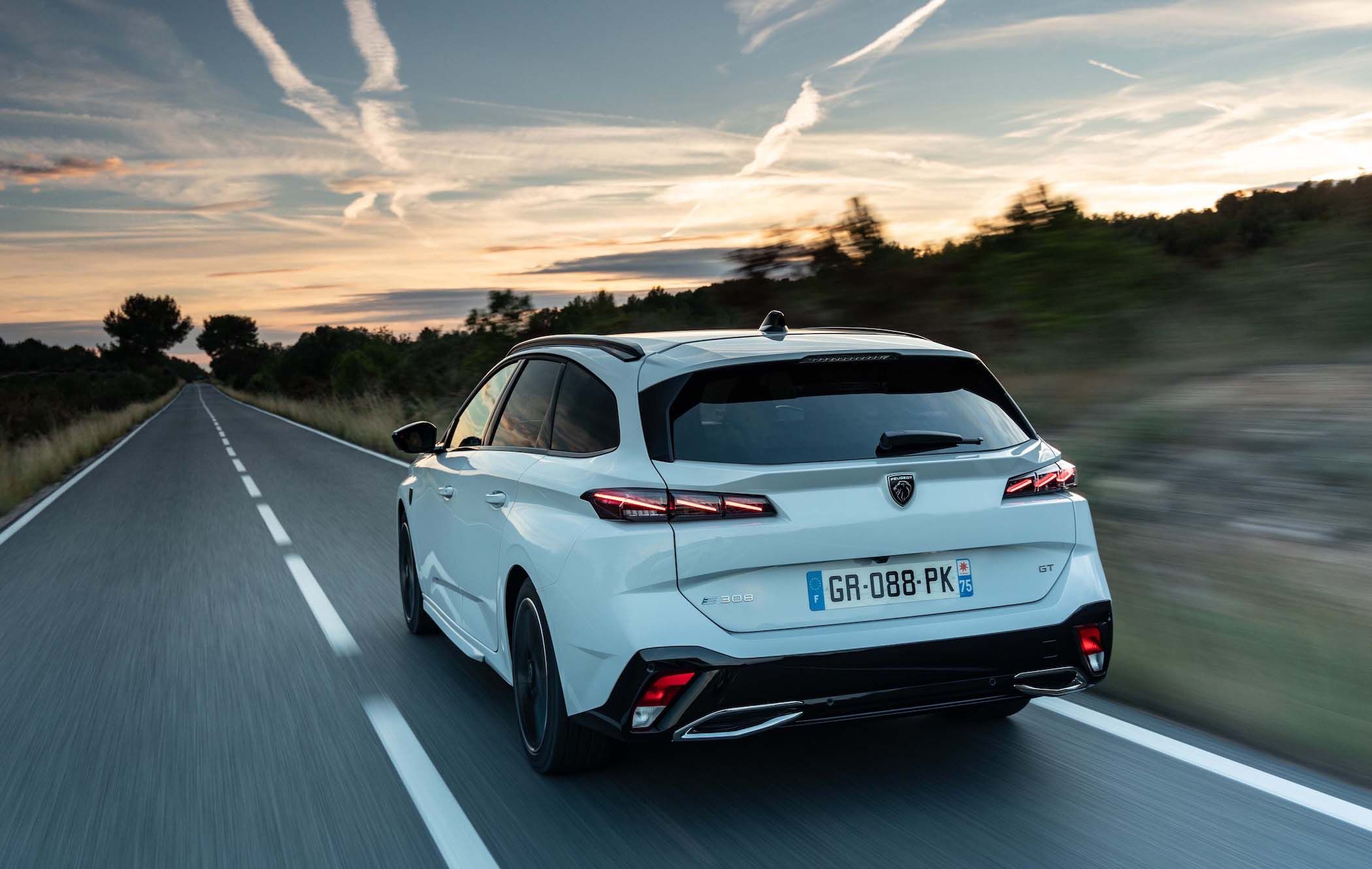 A white Peugeot e-308 SW drives down a rural road at sunset, with trees lining the road and a sky filled with scattered clouds.