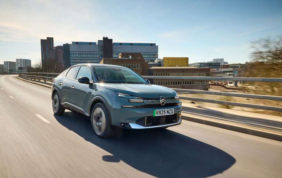 A Citroën ë-C4 X drives on an urban highway with buildings in the background under a clear blue sky.