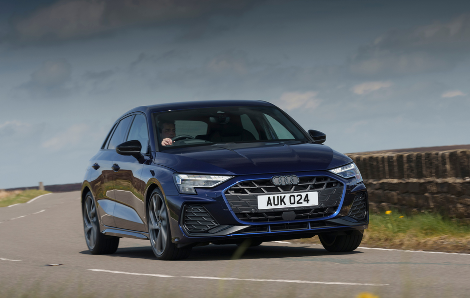 A blue Audi A3 drives on a rural road with stone walls and a cloudy sky in the background.