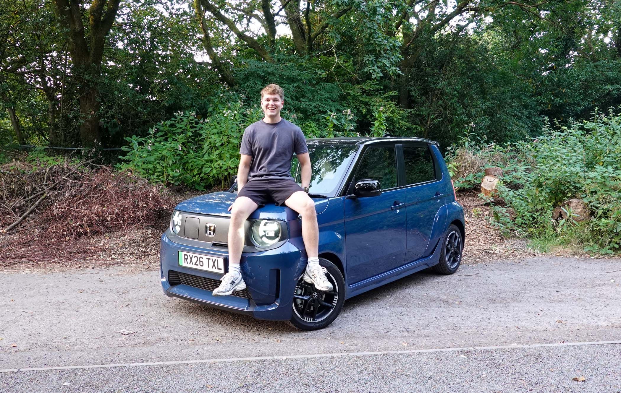 Craig Hale sitting on the bonnet of a blue Honda Super-N parked by a wooded area.