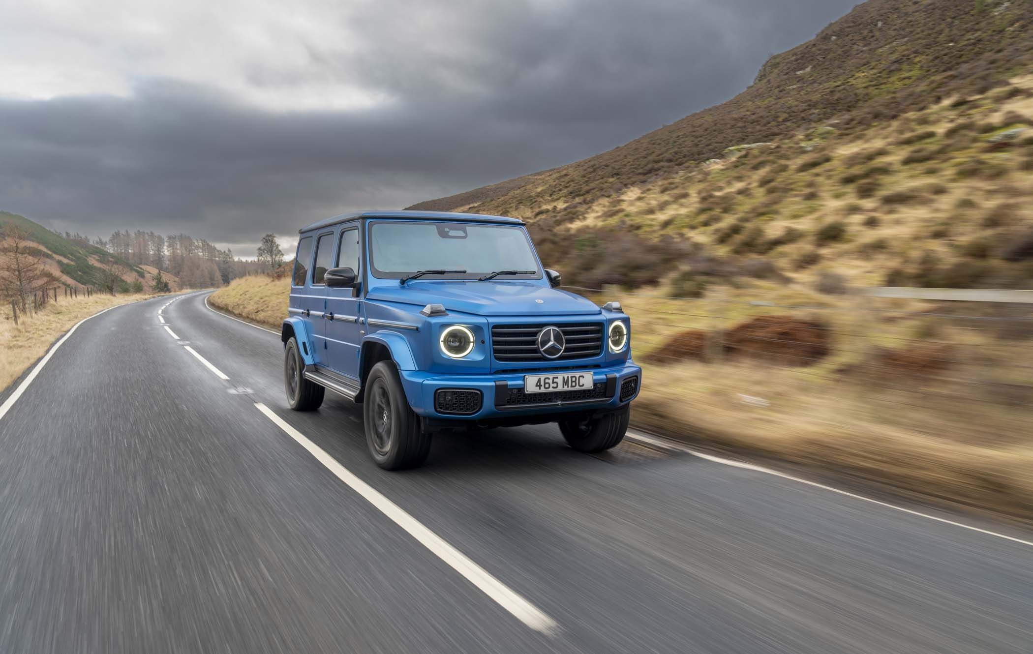 A blue Mercedes G-Class with EQ Technology drives on a winding rural road, surrounded by hills and cloudy skies.