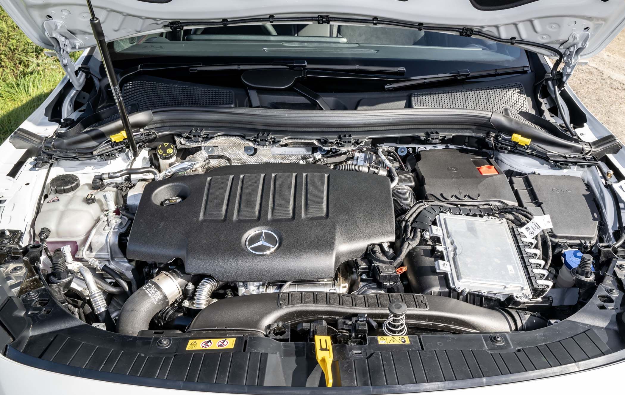 Mercedes GLA car engine with a black cover featuring the Mercedes logo, surrounded by various engine components and a white exterior.