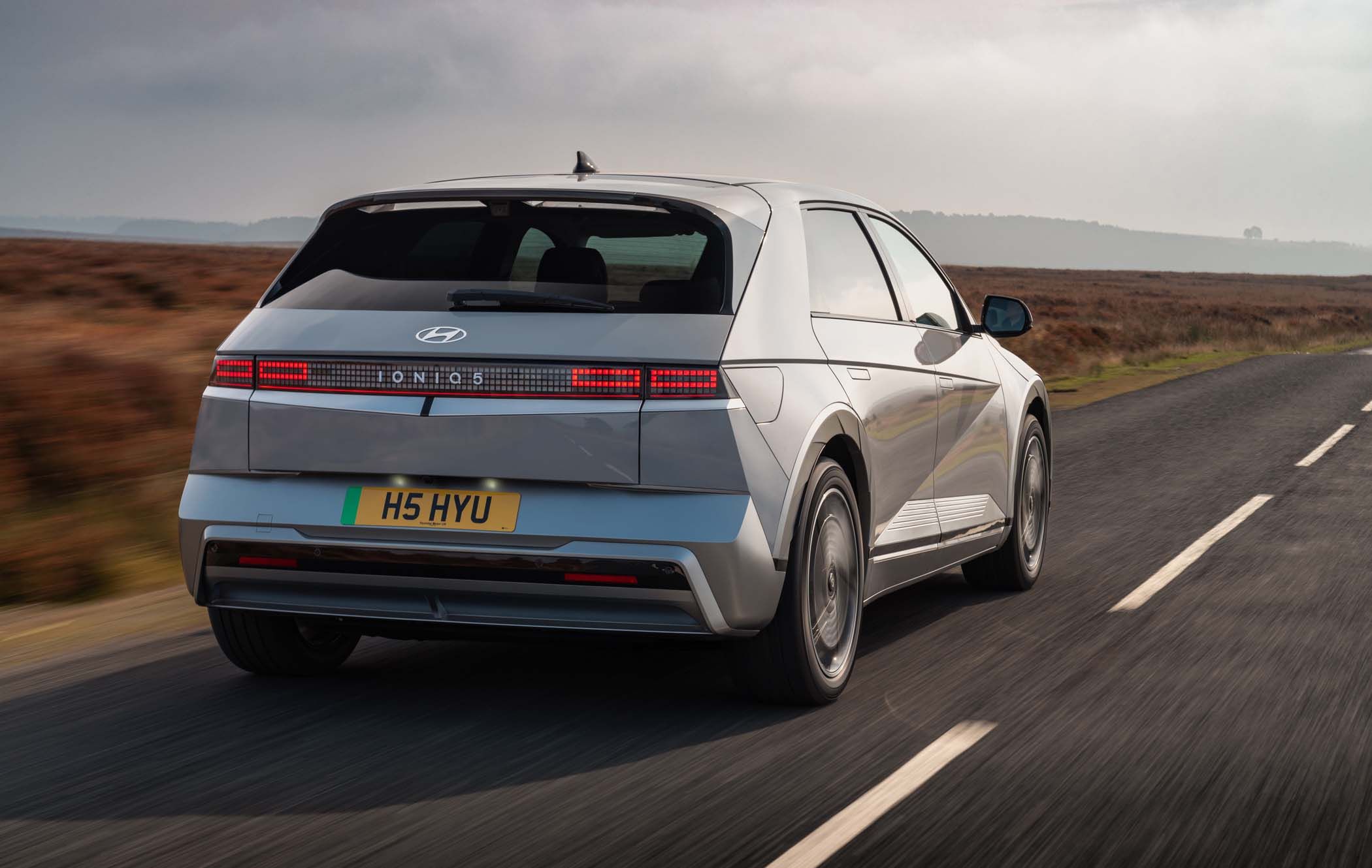 A silver Hyundai IONIQ 5 electric car drives on a rural road under a cloudy sky, showcasing its modern design and rear view.
