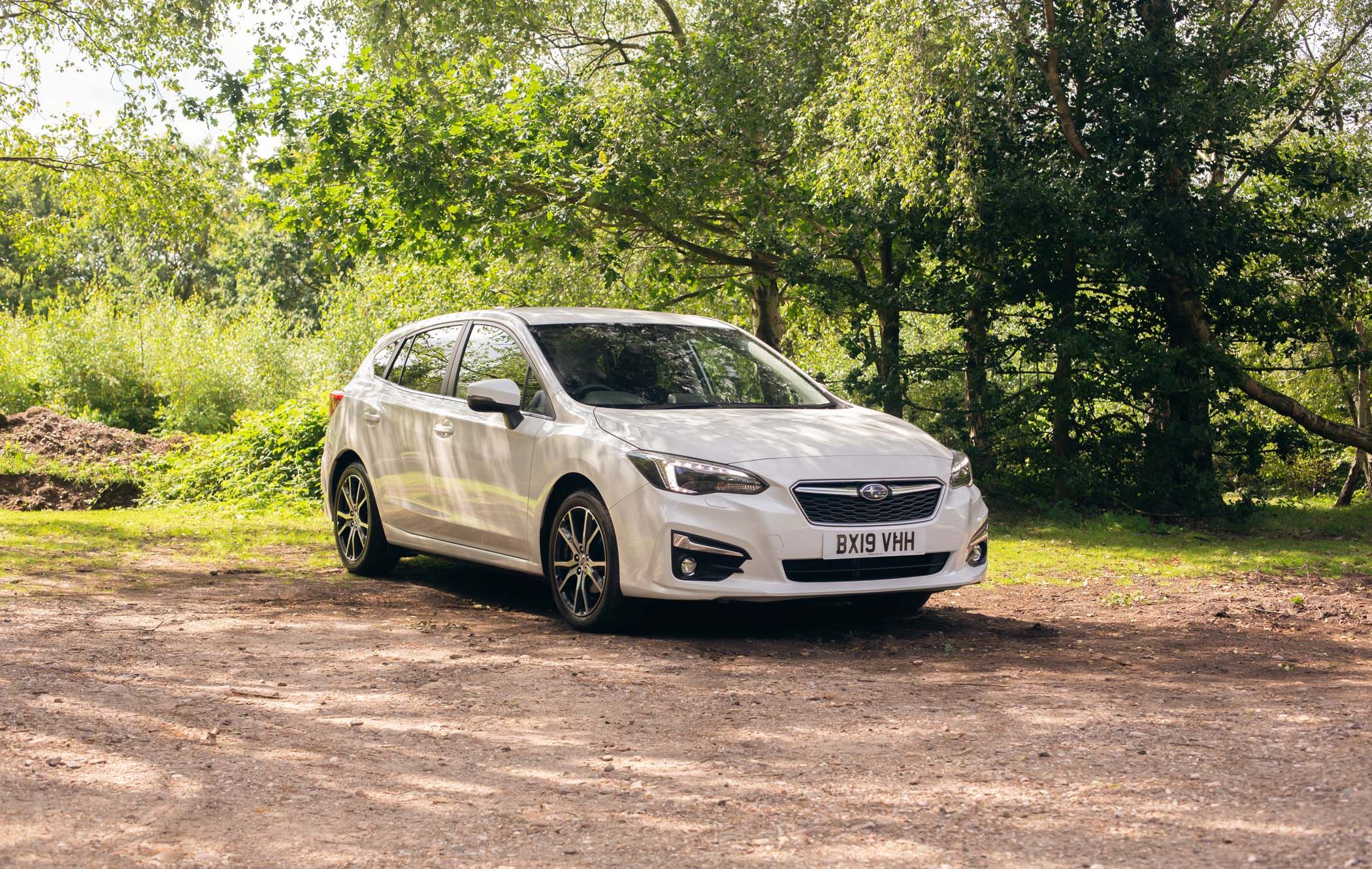 A white Subaru Impreza parked on a dirt path surrounded by lush green trees and sunlight filtering through the leaves.