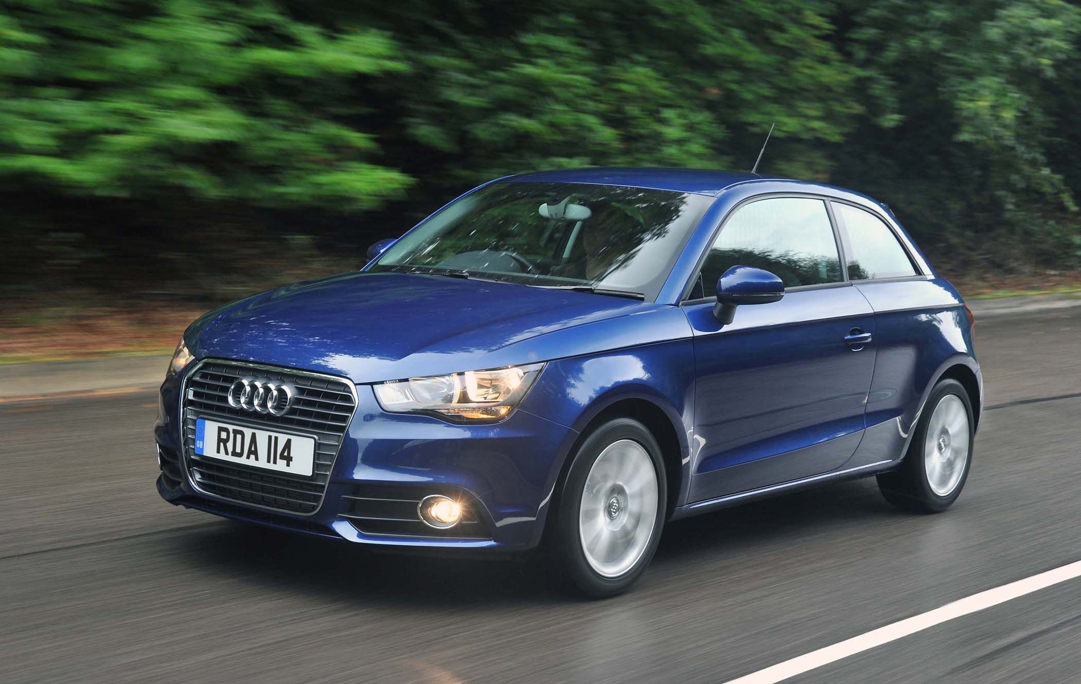 A blue Audi A1 driving on a wet road with green trees in the background.
