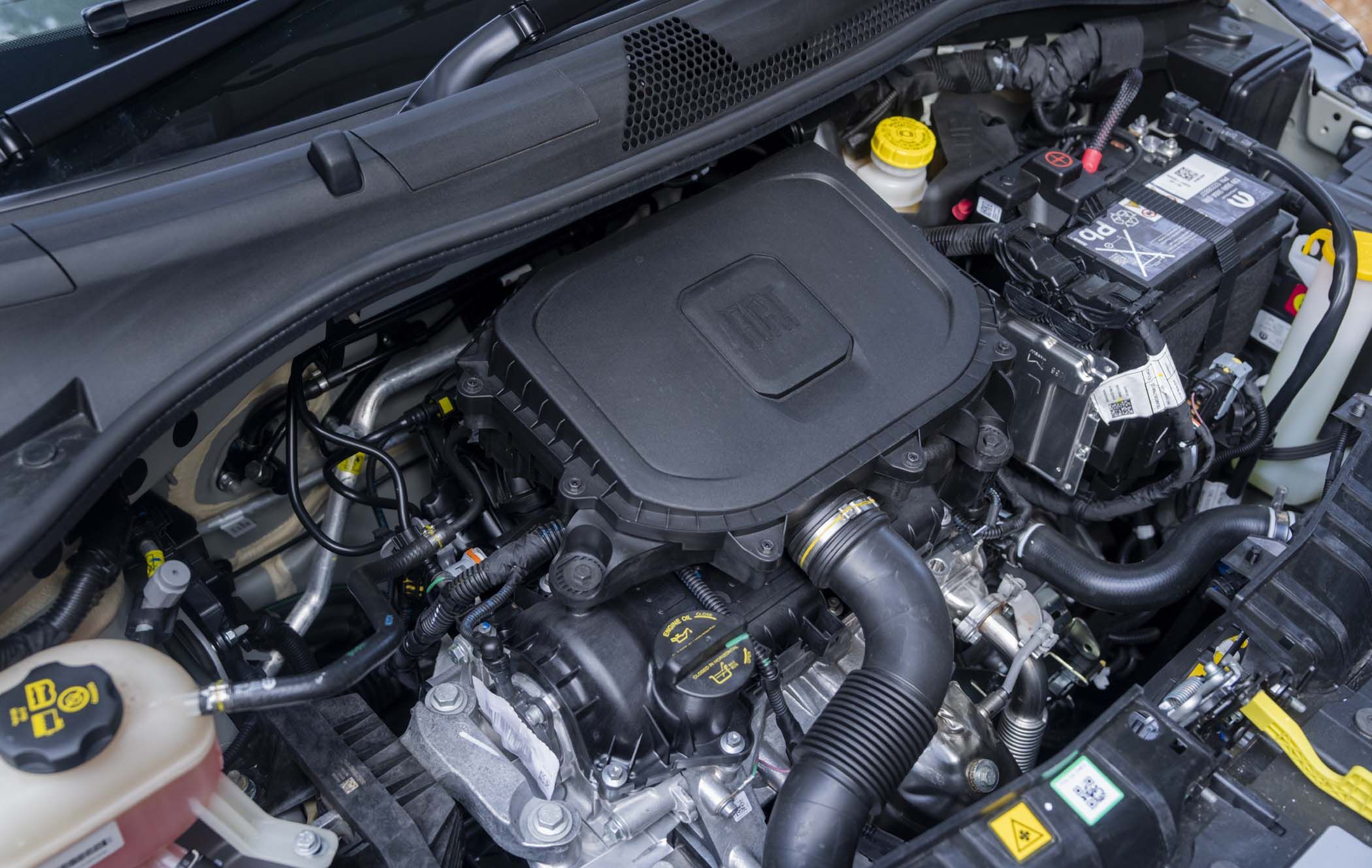 A close-up of a Fiat 500 Hybrid engine compartment showing various components, including the battery, air intake, hoses, and reservoirs.