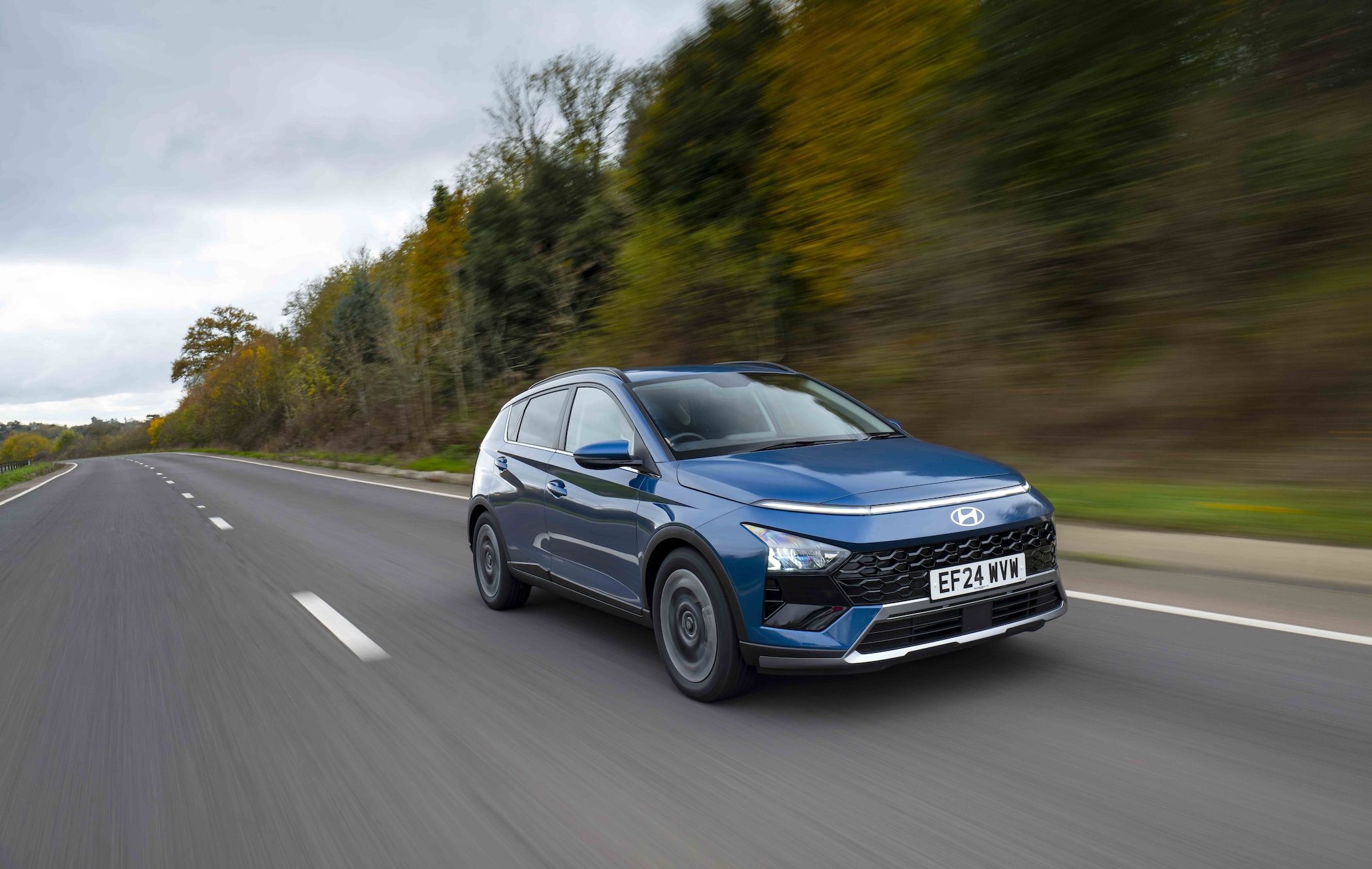 A blue Hyundai Bayon driving on a highway with blurred trees in the background under a cloudy sky.