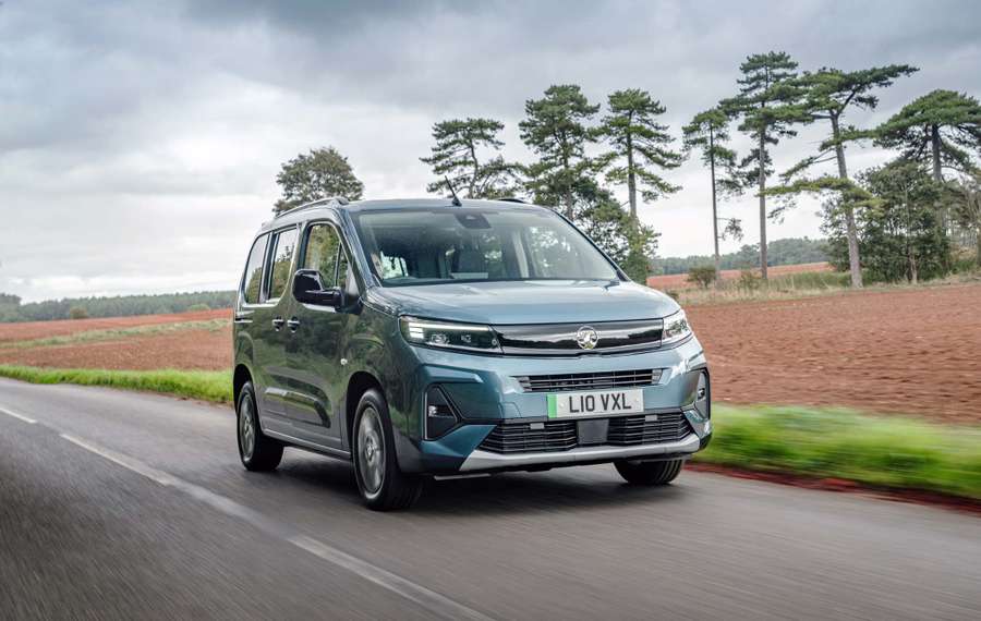 A blue Vauxhall Combo Life Electric driving on a rural road with trees and fields in the background under a cloudy sky.