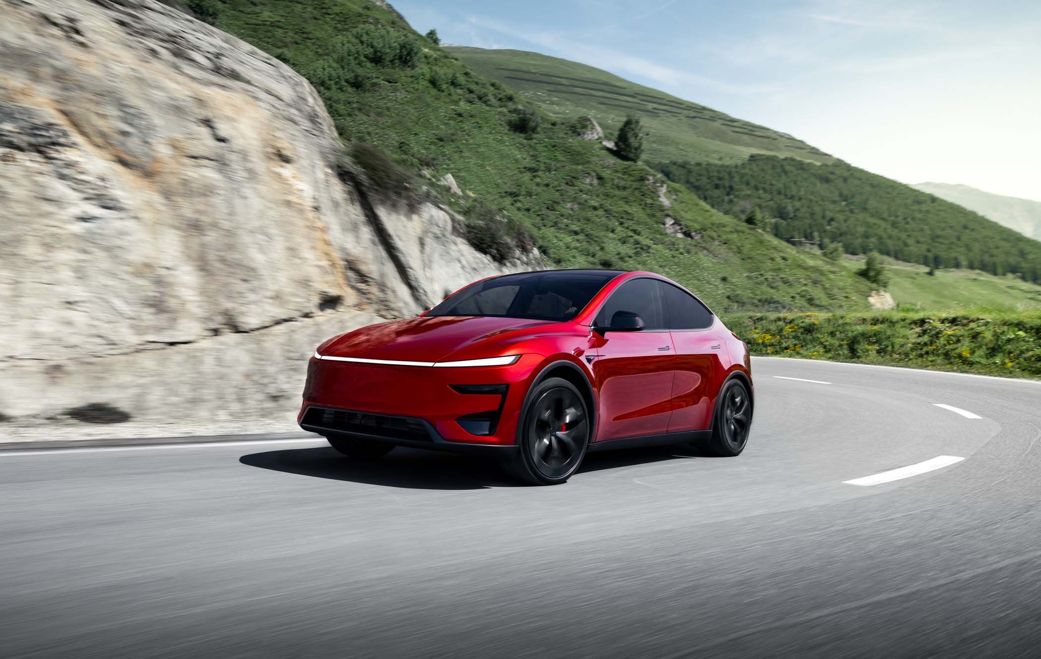 A red Tesla Model Y Performance drives on a winding mountain road, with lush green hills and a clear blue sky in the background.