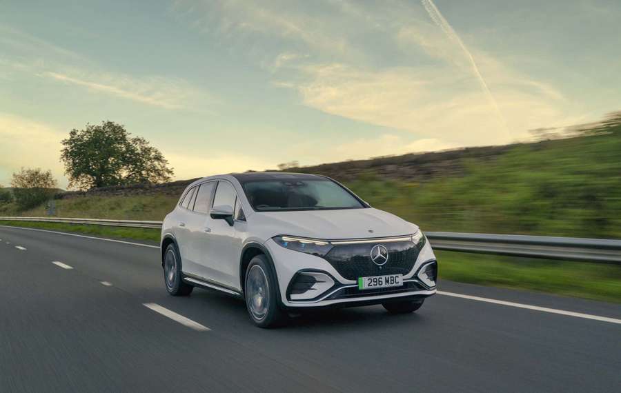 A white Mercedes EQS SUV driving on a highway with a grassy landscape and a tree in the background under a partly cloudy sky.