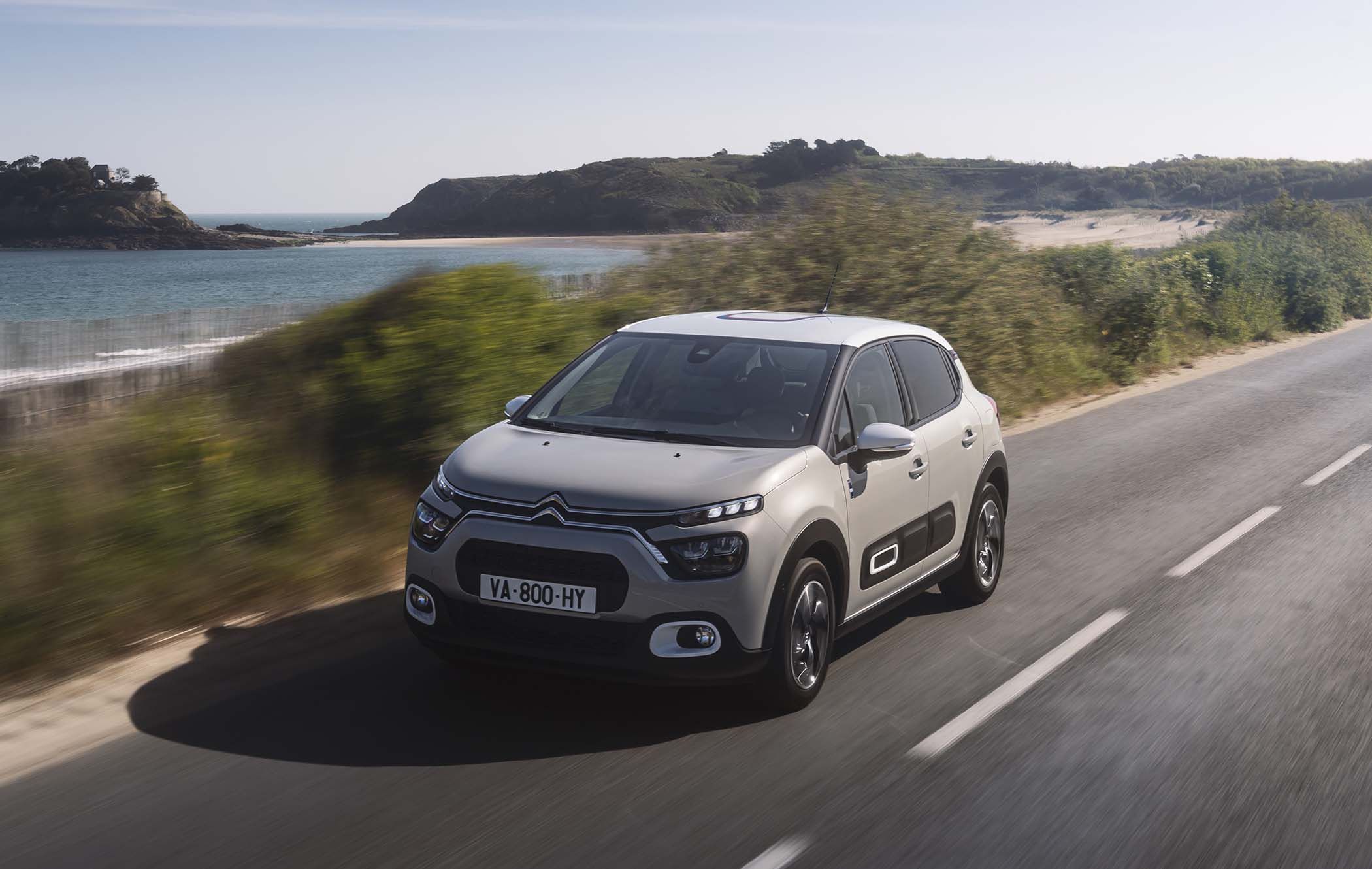 Grey Citroën C3 driving on a coastal road with greenery and ocean in the background under a clear sky.
