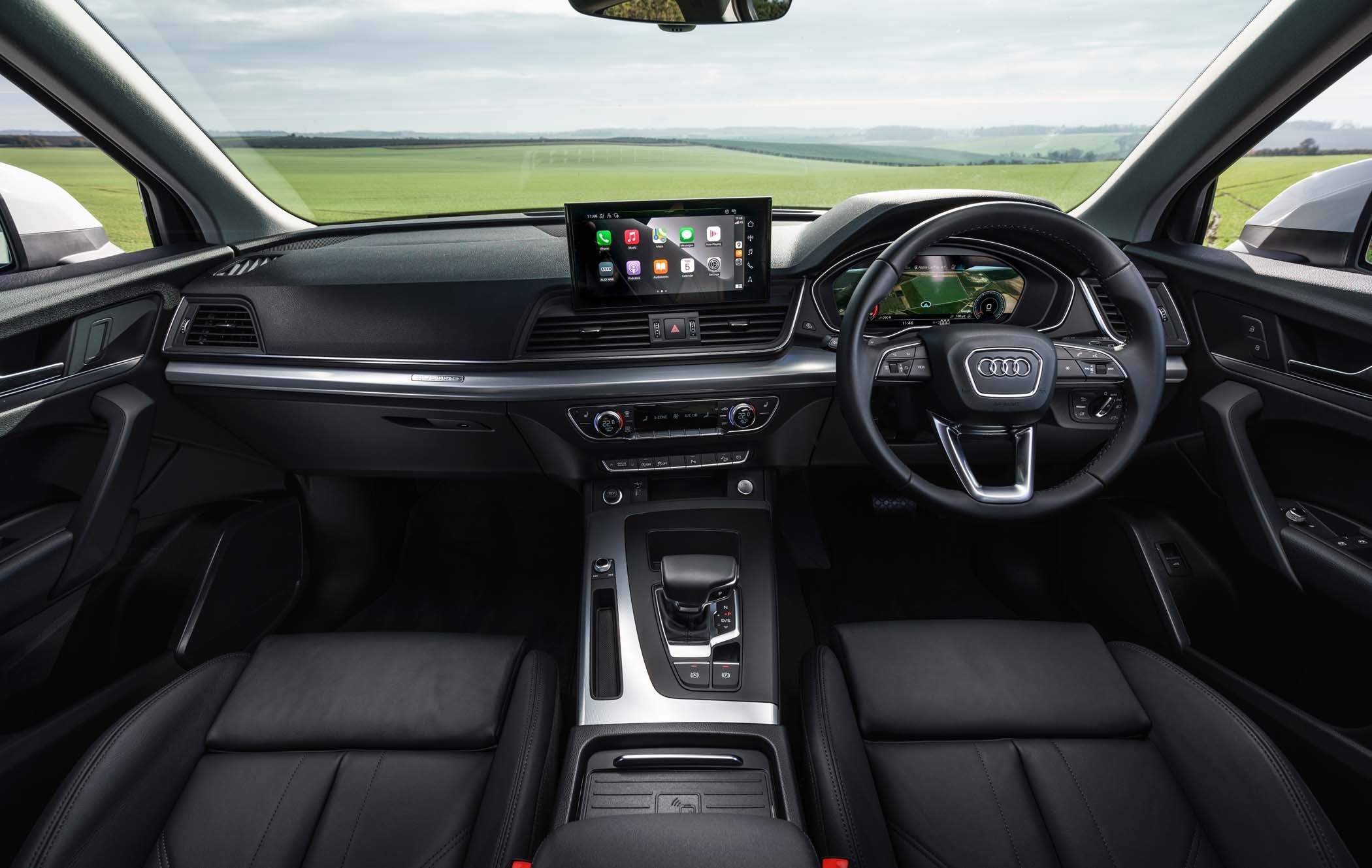 Interior view of an Audi Q5 featuring a black leather dashboard, steering wheel, touchscreen display, gear shift, and front seats against a rural backdrop.