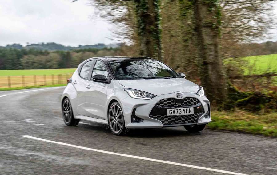 A silver Toyota Yaris car drives on a winding road lined with trees and fields under a cloudy sky.