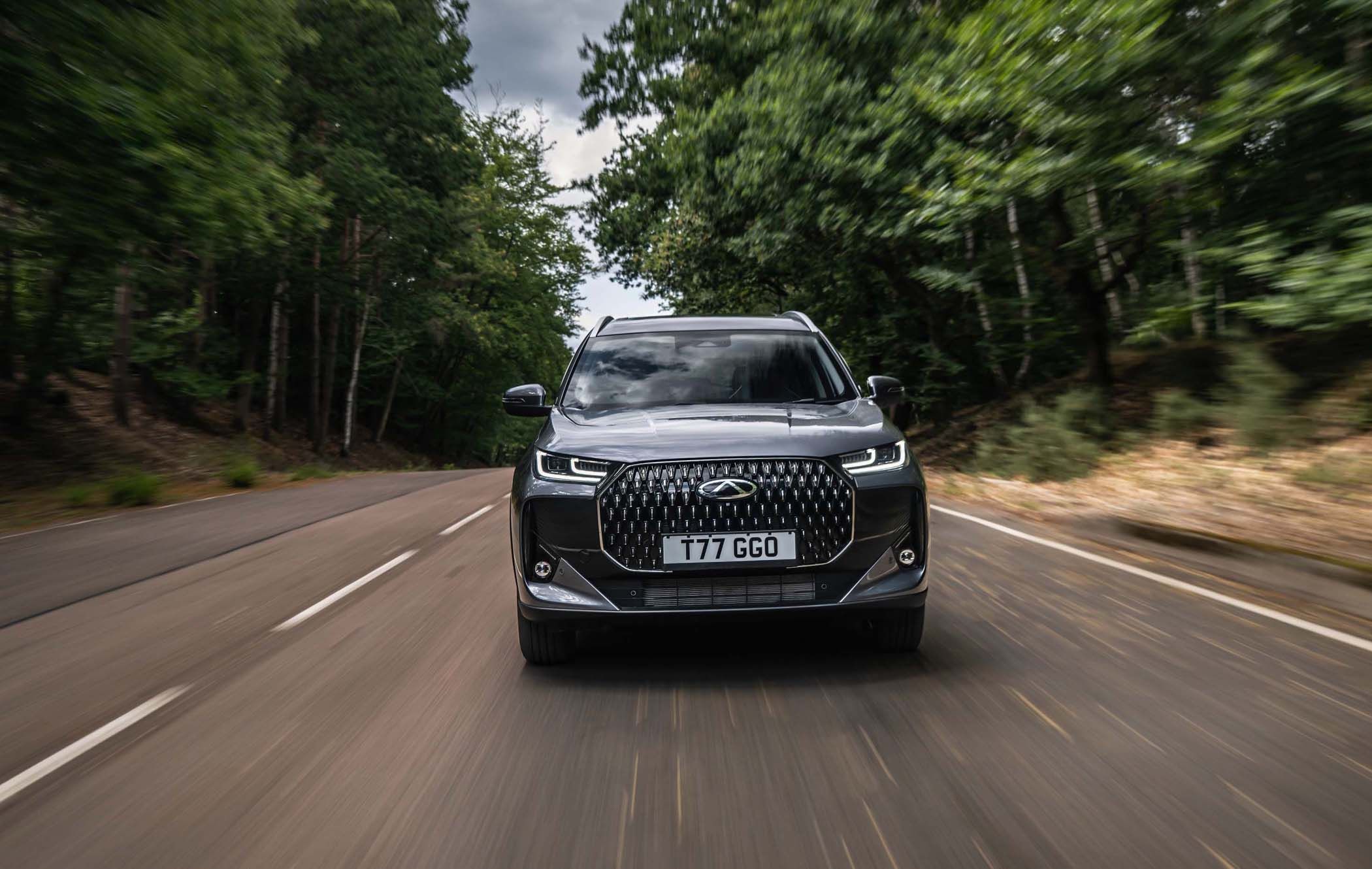 A Chery Tiggo 7 Super Hybrid drives on a forest road, surrounded by blurred green trees, under a cloudy sky.