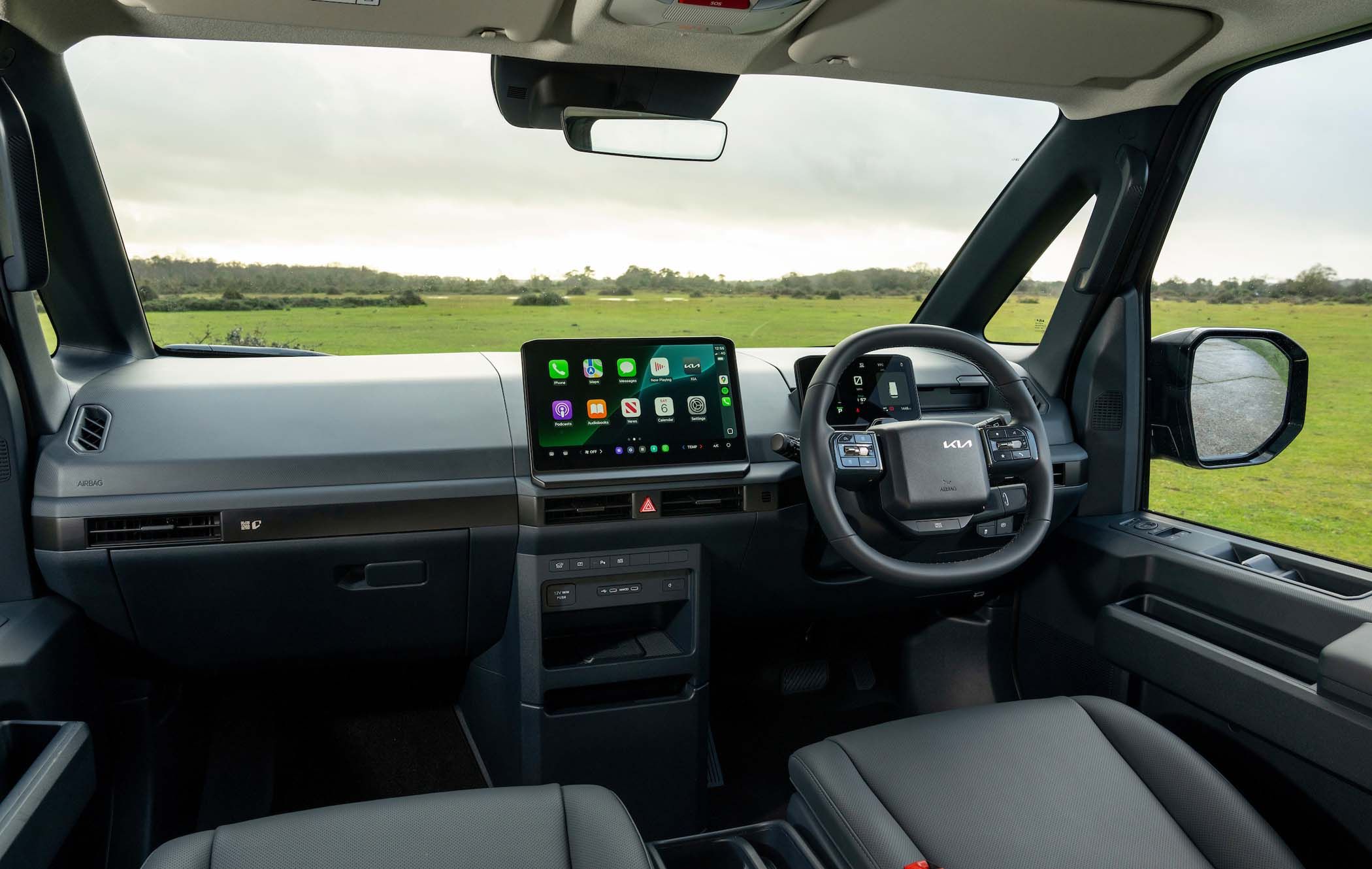 Interior of a Kia PV5 Passenger with a digital dashboard, touchscreen displaying apps, steering wheel, and leather seats, overlooking a grassy field.