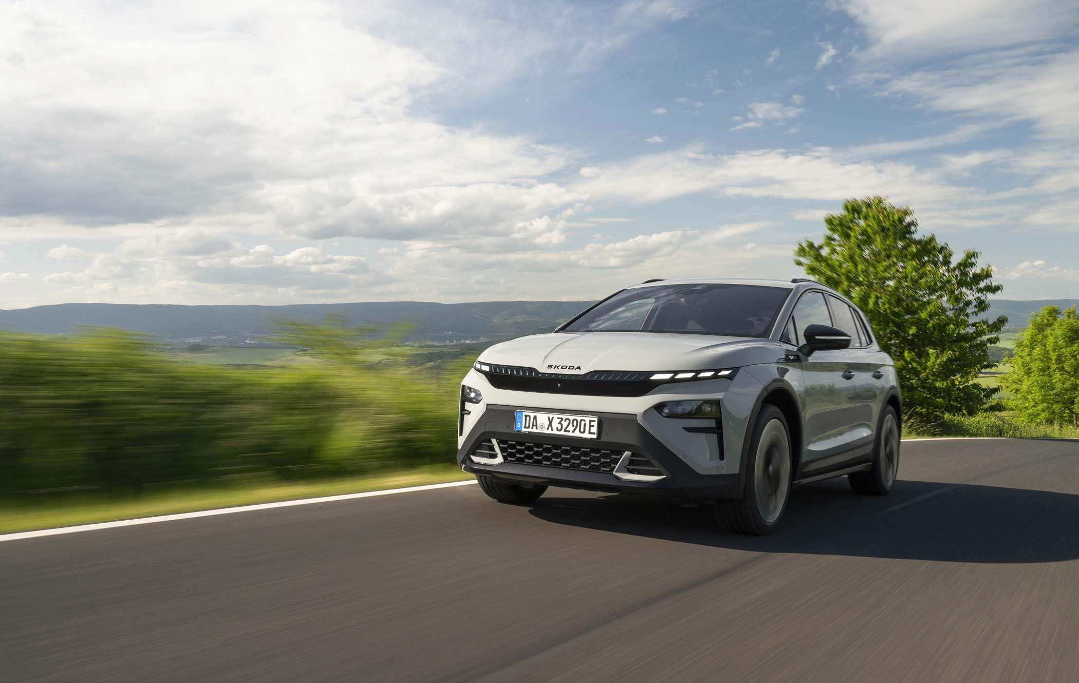 A silver Škoda Elroq drives on a scenic road with lush greenery and hills under a partly cloudy sky.