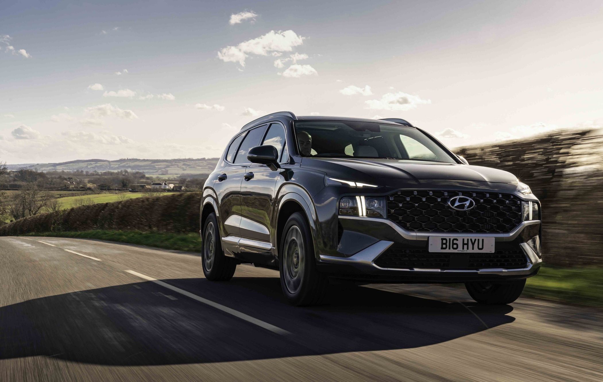 A black Hyundai Santa Fe driving on a rural road with fields and hills in the background under a partly cloudy sky.